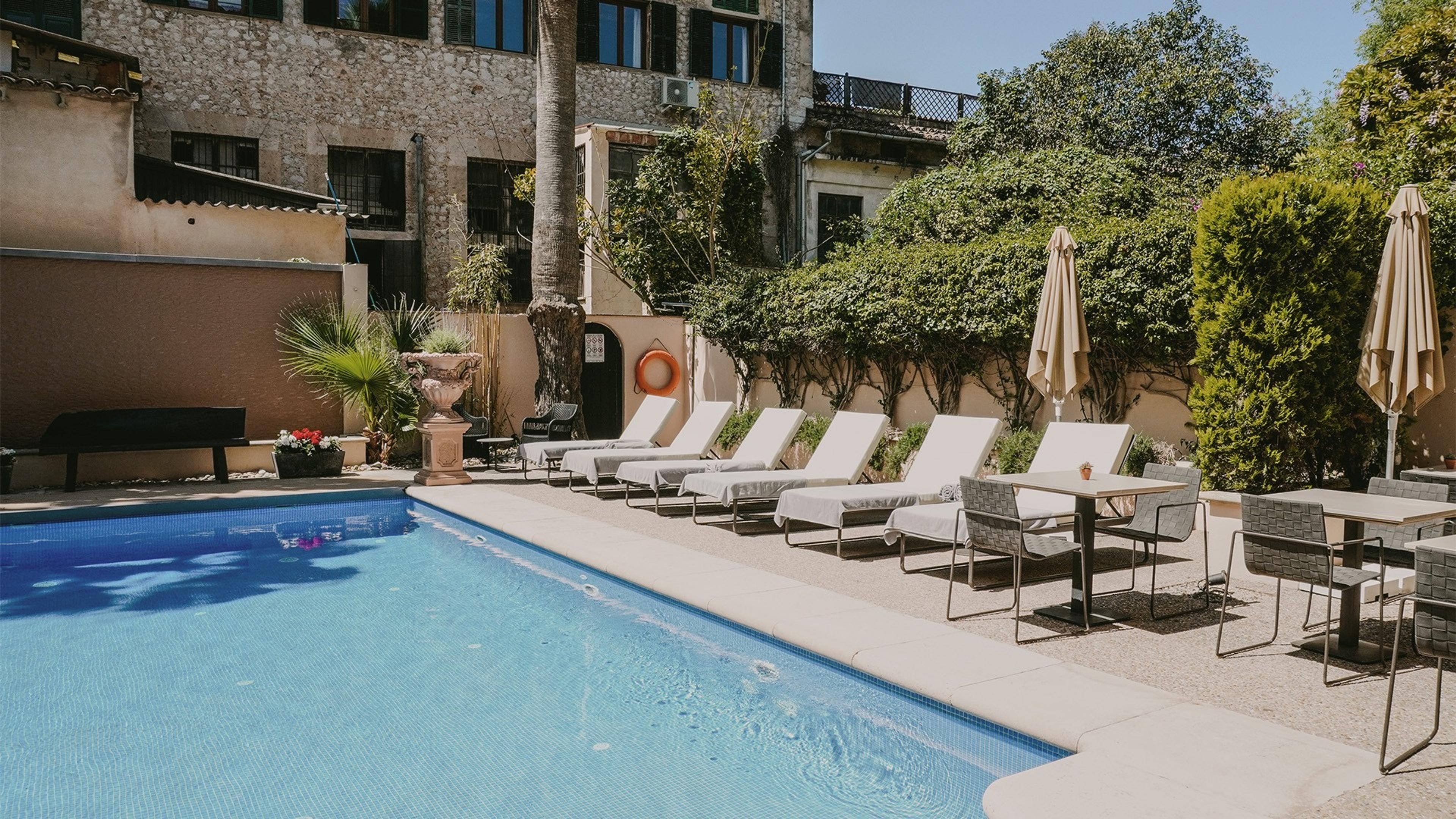 An outdoor swimming pool beside sun loungers and shaded tables, with an old stone building, a palm tree, and trimmed hedges behind.