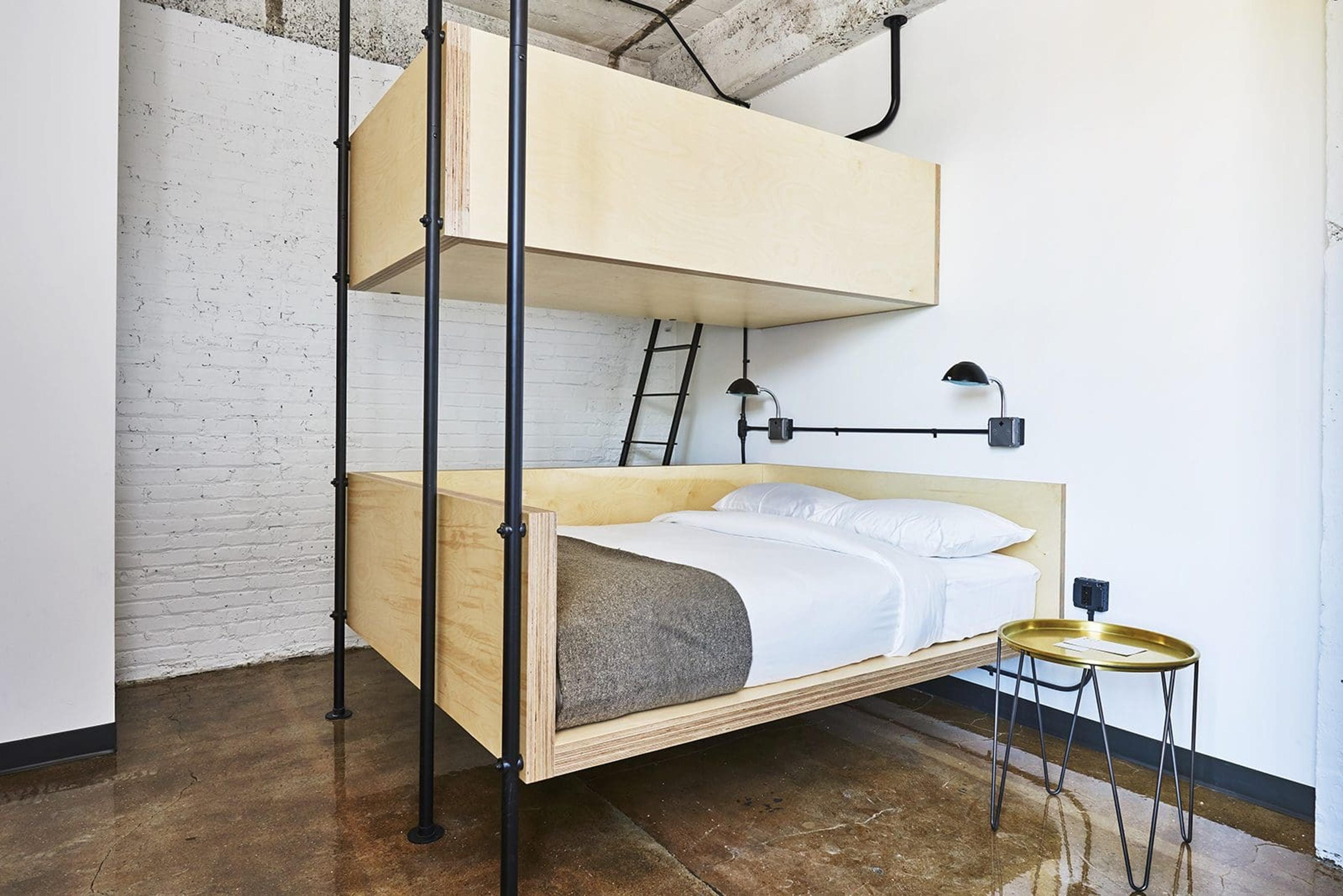 a plywood bunk bed with a ladder and black metal frame, the lower bed made up with white linens and a grey throw, under a wall-mounted reading lamp and exposed concrete and brick walls