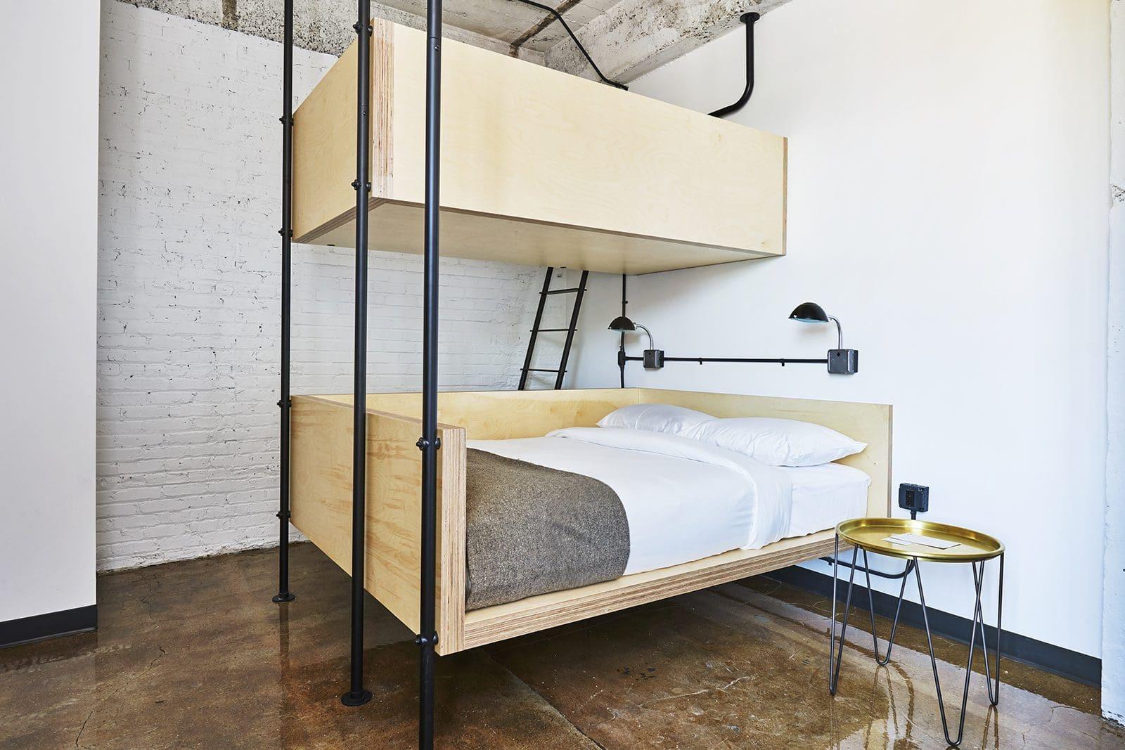 a plywood bunk bed with a ladder and black metal frame, the lower bed made up with white linens and a grey throw, under a wall-mounted reading lamp and exposed concrete and brick walls