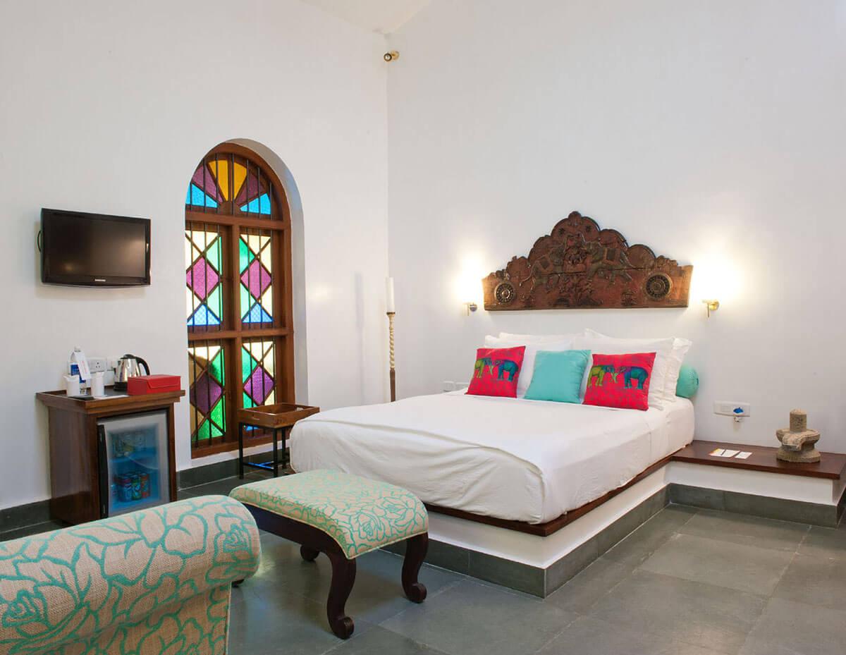 A bedroom with a carved wooden headboard, red and teal cushions and a stained glass arched window beside a television and minibar.