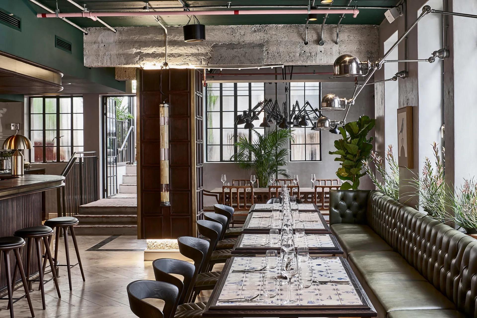A dining room with a green leather banquette along one wall, long tables with glasses and blue-patterned tile tops, and bar stools and metal pendant lamps overhead.