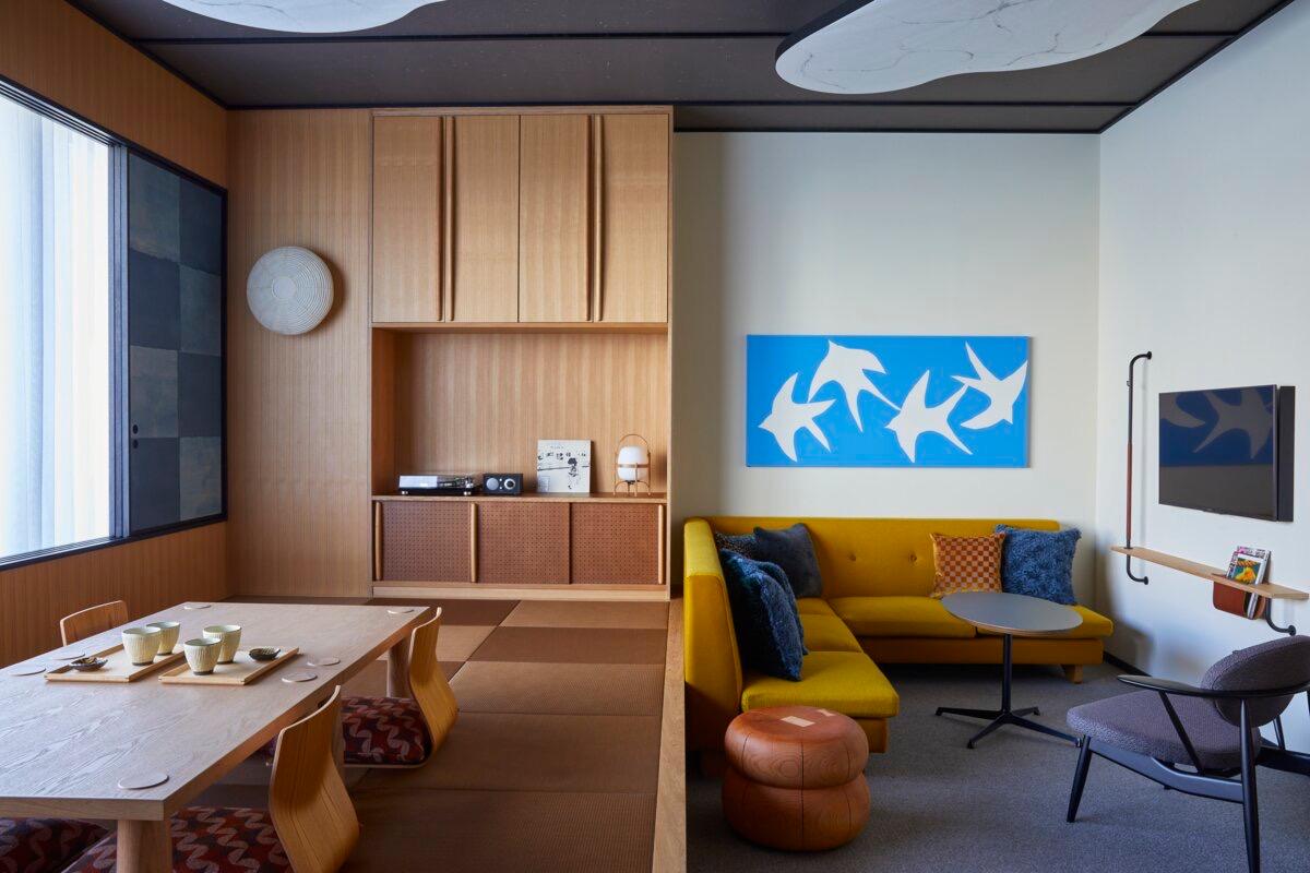 A hotel suite combining a low tatami dining table with floor cushions and a built in wood cabinet on one side, and a mustard yellow sectional sofa under a blue swallow print on the other.