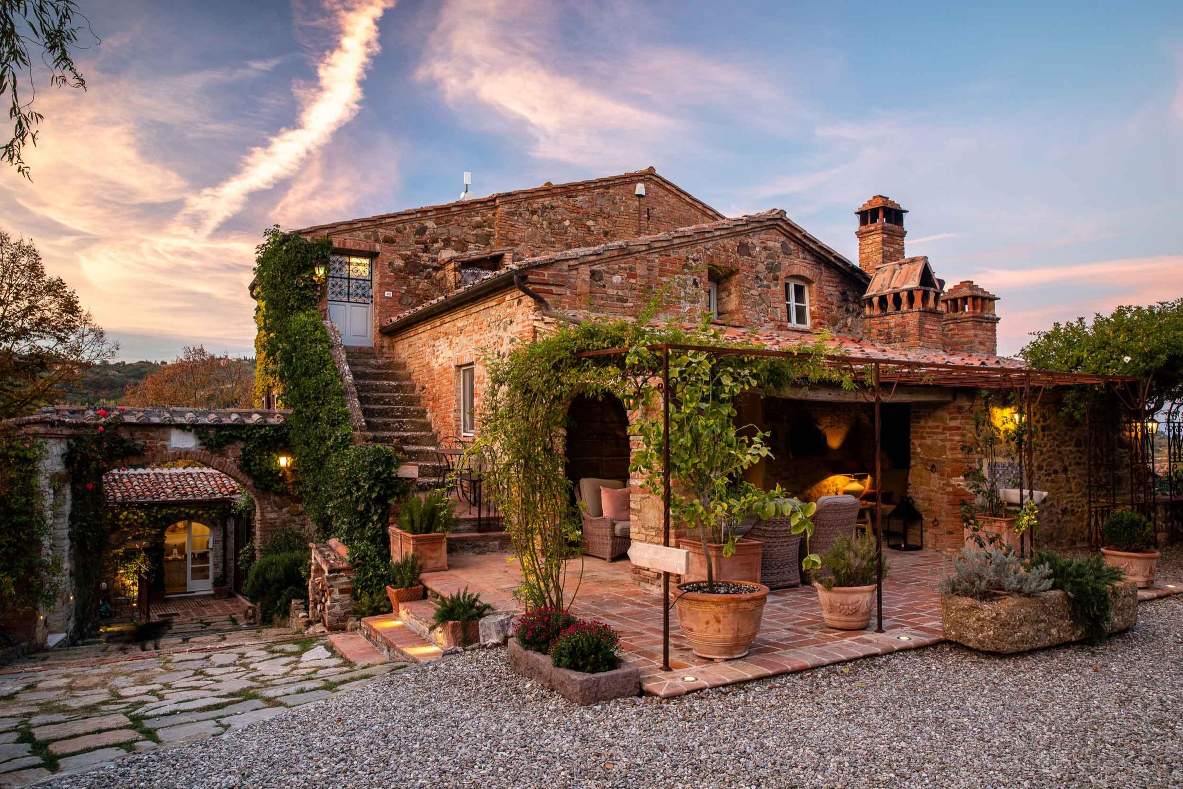 A stone farmhouse at sunset, with a covered terrace, potted plants and lit windows, under a pink and blue sky.