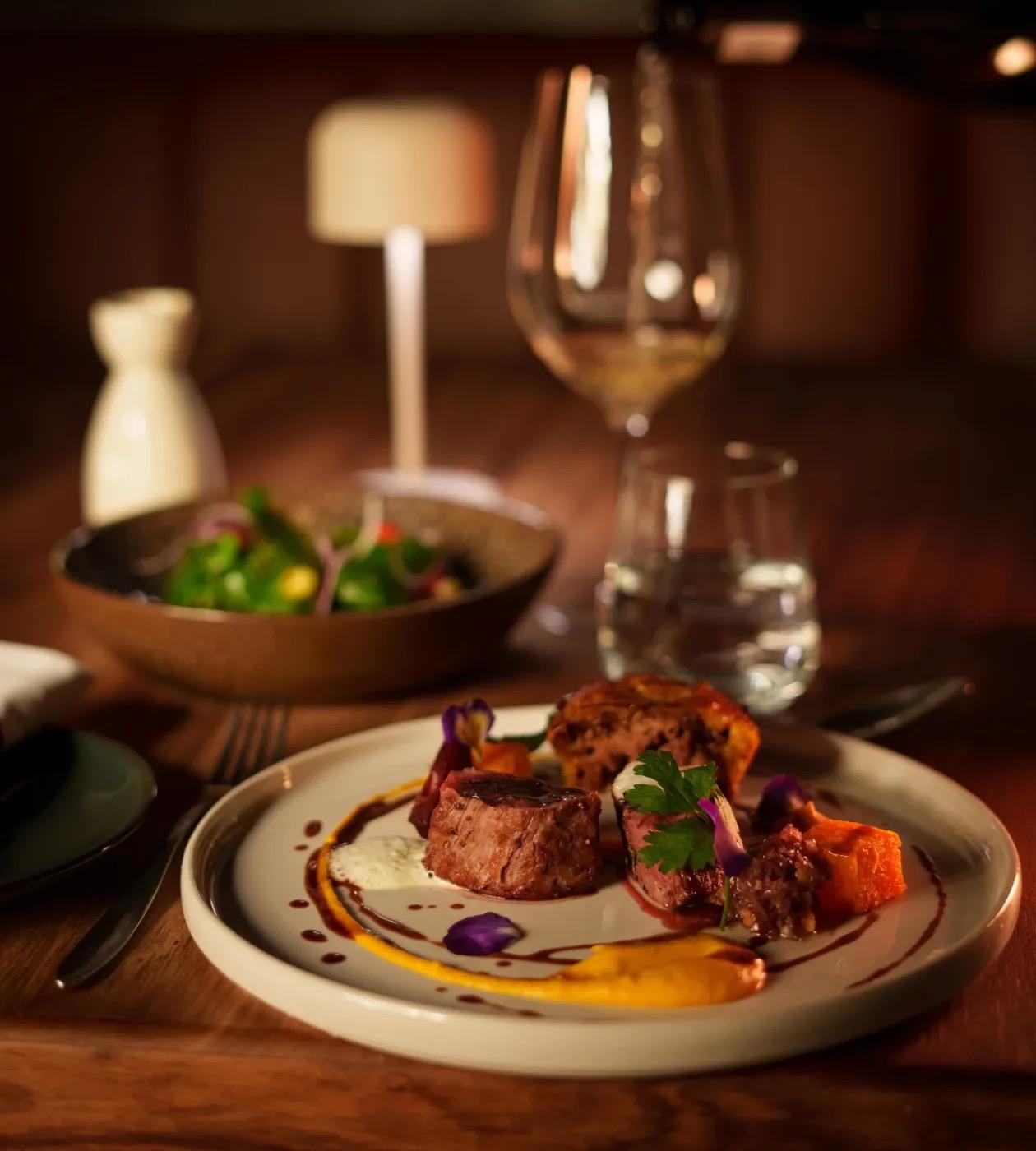 Two medallions of seared beef on a plate with a golden purée, roasted squash and a small salad, beside a glass of white wine.