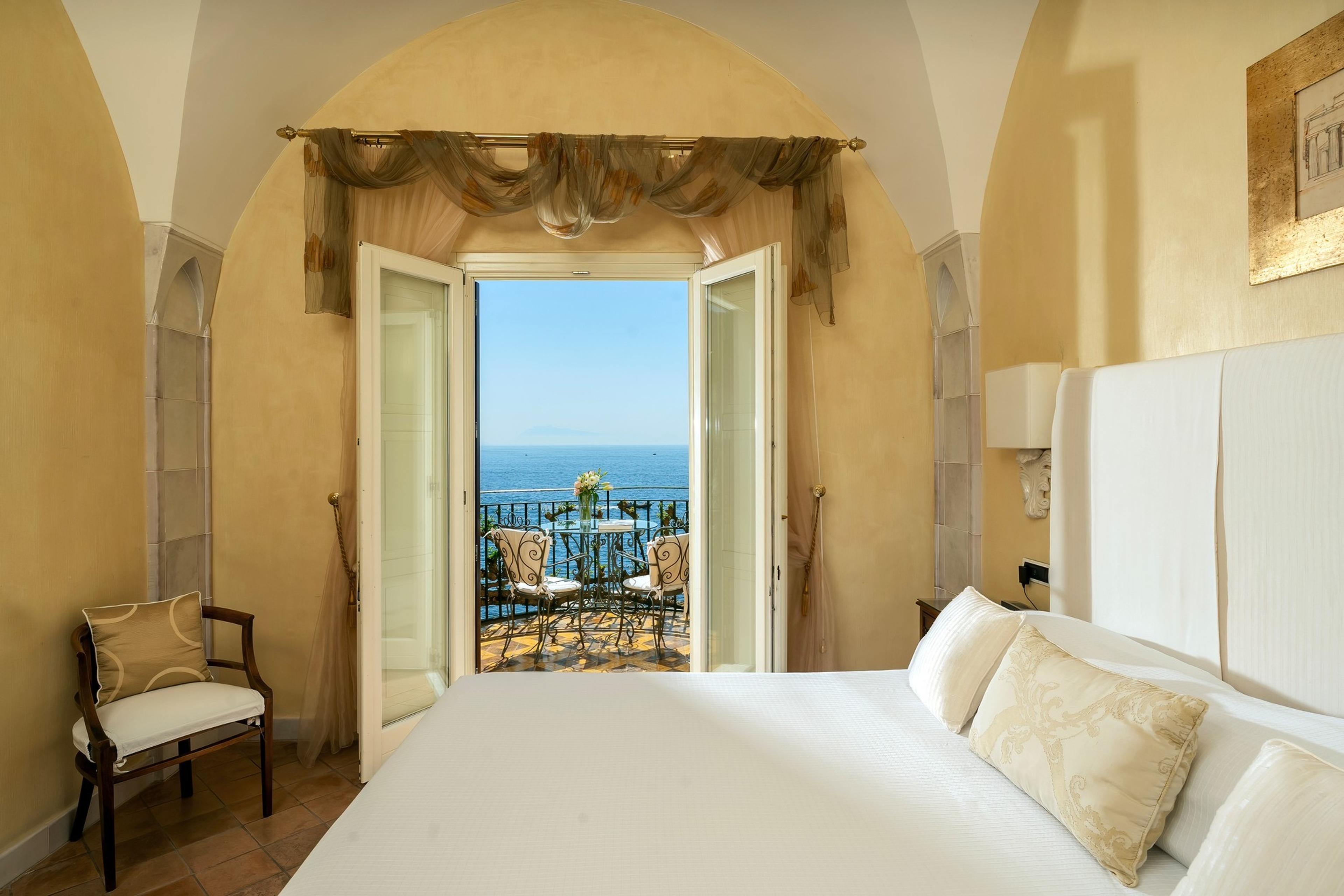 A bedroom under a vaulted ceiling, with a bed facing an arched doorway open to a small balcony with a table, chairs and a sea view.