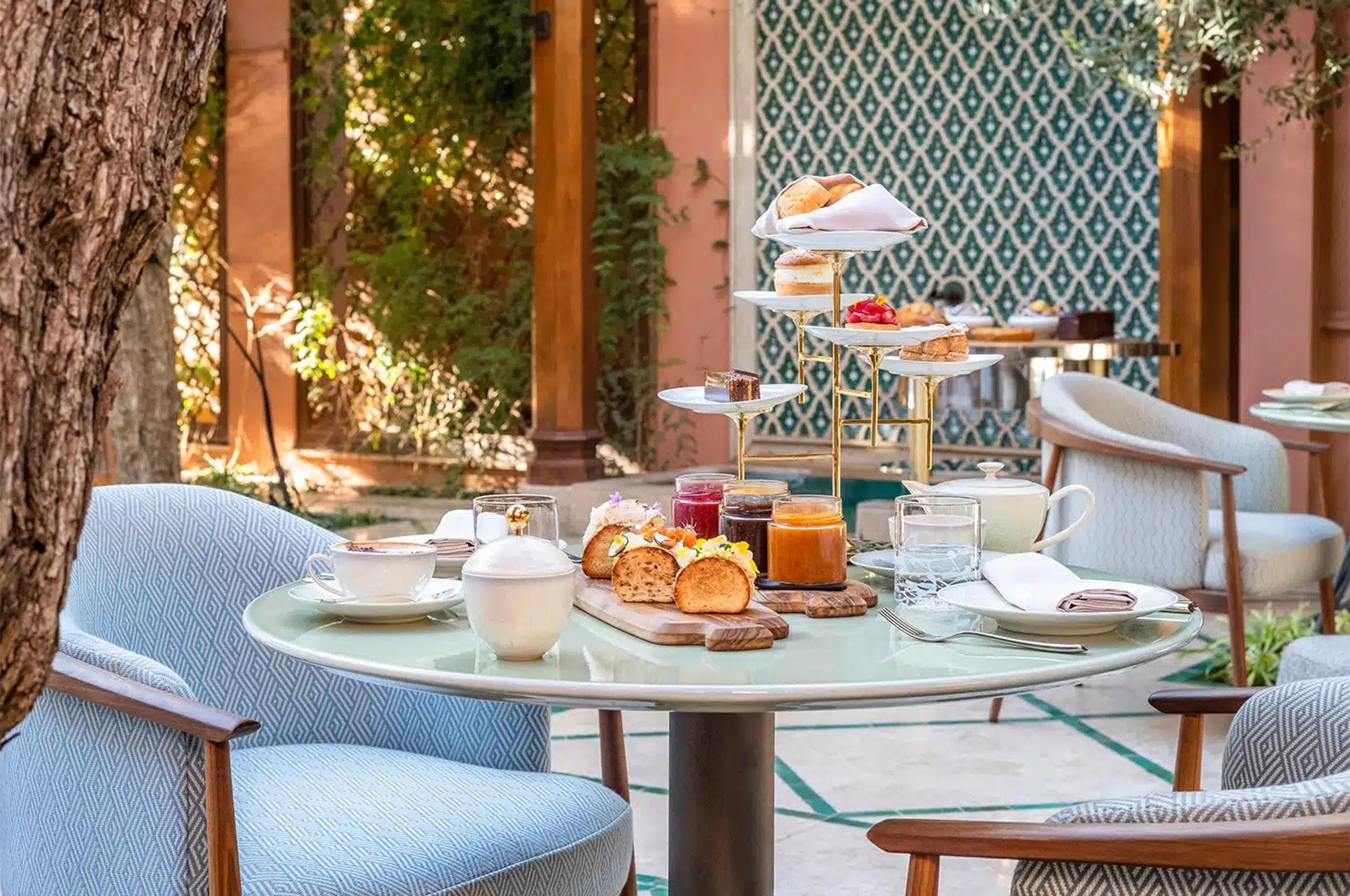 A round outdoor table set with tea, pastries, jam and a tiered cake stand beside a patterned wall.