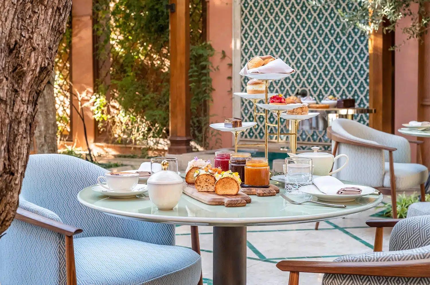 A round outdoor table set with tea, pastries, jam and a tiered cake stand beside a patterned wall.