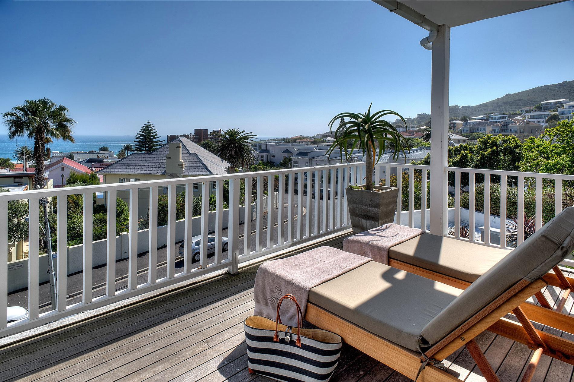 A timber balcony with two sunloungers, looking over rooftops and a palm tree to a stretch of sea under a clear sky.