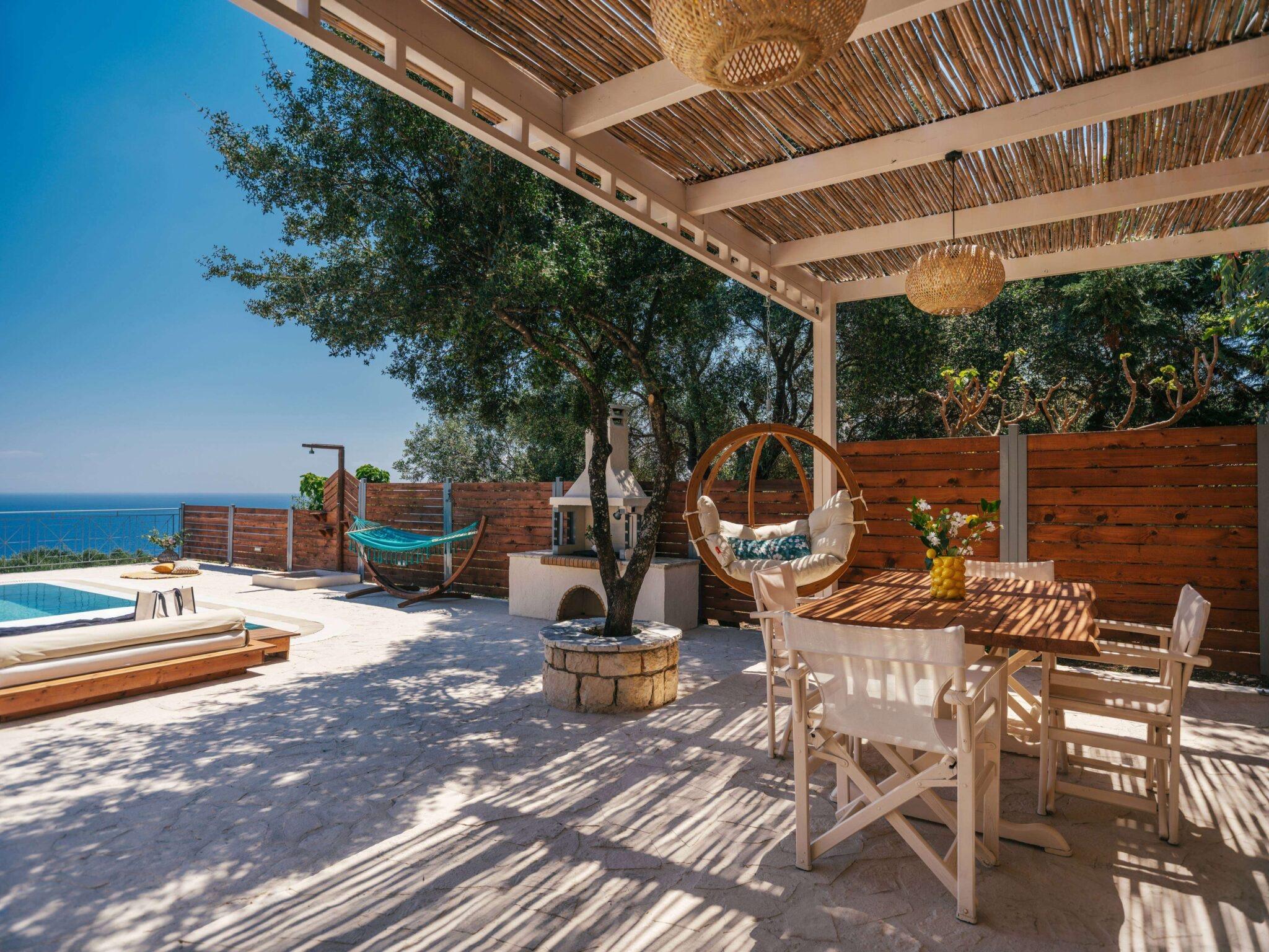a covered outdoor dining terrace with a wooden table set for four, a hanging egg chair, a hammock and a stone barbecue, olive trees behind