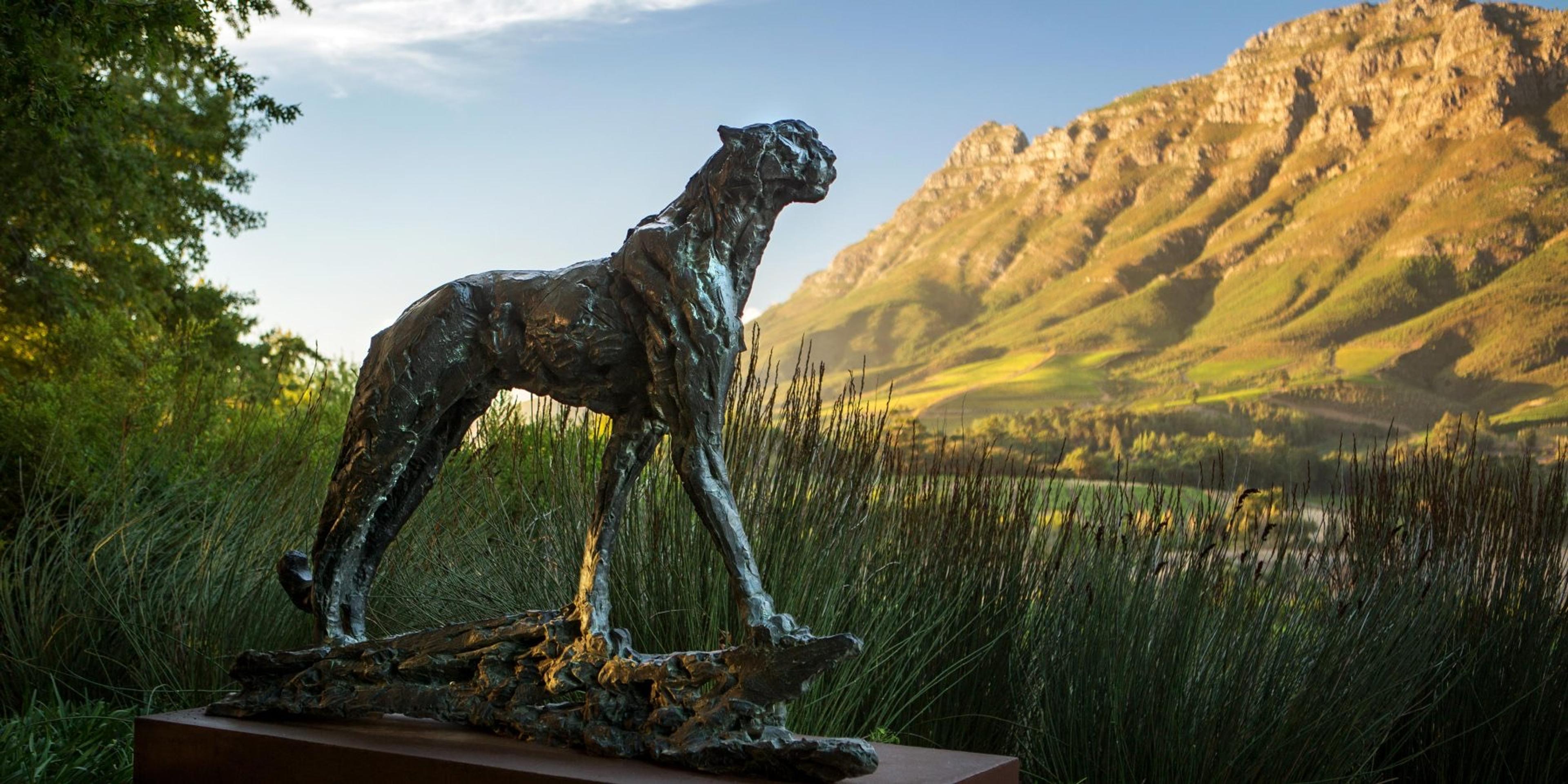 A bronze cheetah sculpture stands on driftwood among tall grass, a mountain range behind it.