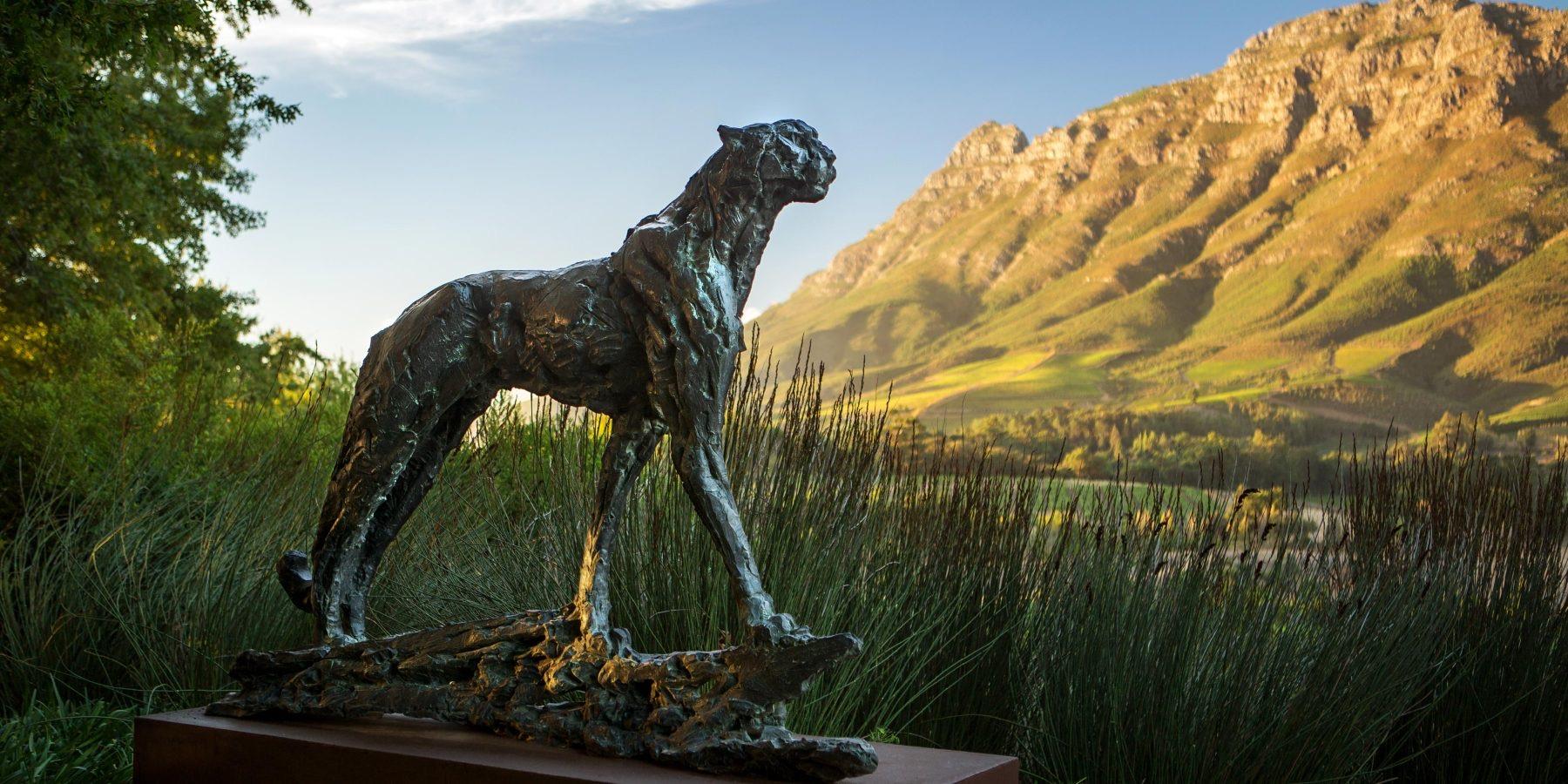 A bronze cheetah sculpture stands on driftwood among tall grass, a mountain range behind it.
