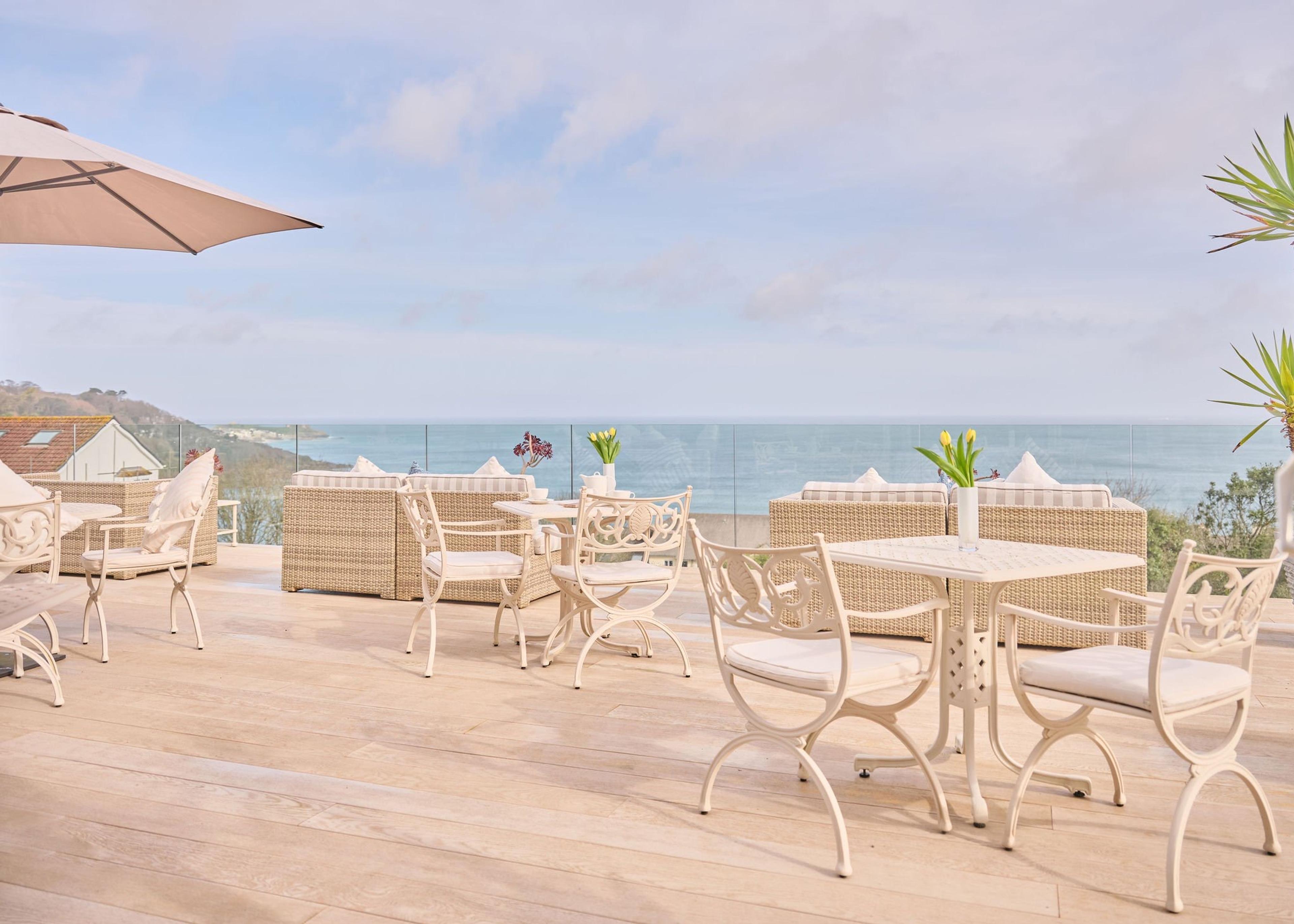 A clifftop terrace with white metal chairs and tables set with flowers, a sun umbrella, and the sea stretching to the horizon under a blue sky.