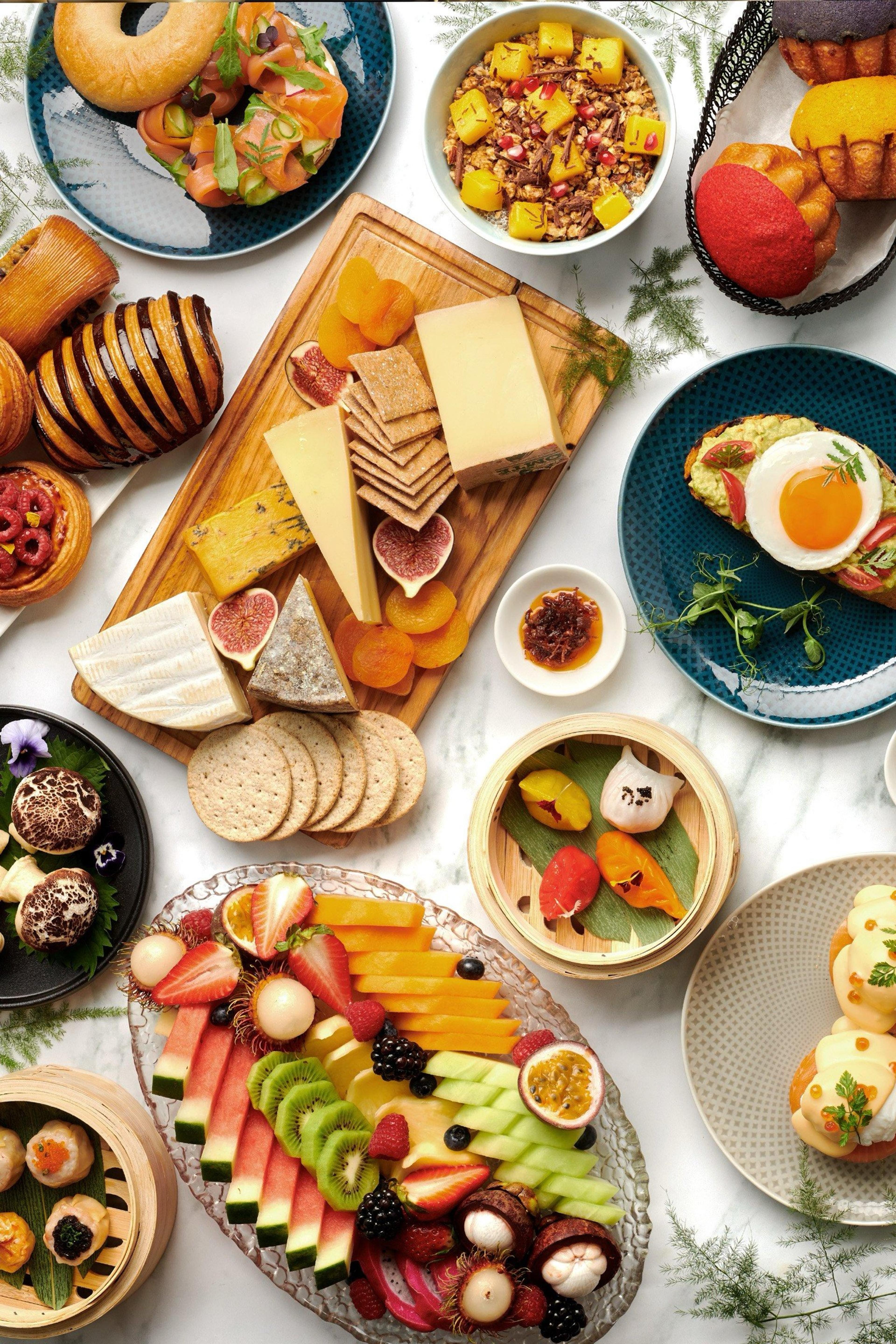 An overhead spread of breakfast dishes including a cheese board, smoked salmon bagel, fruit platter, dim sum and pastries on a marble table.