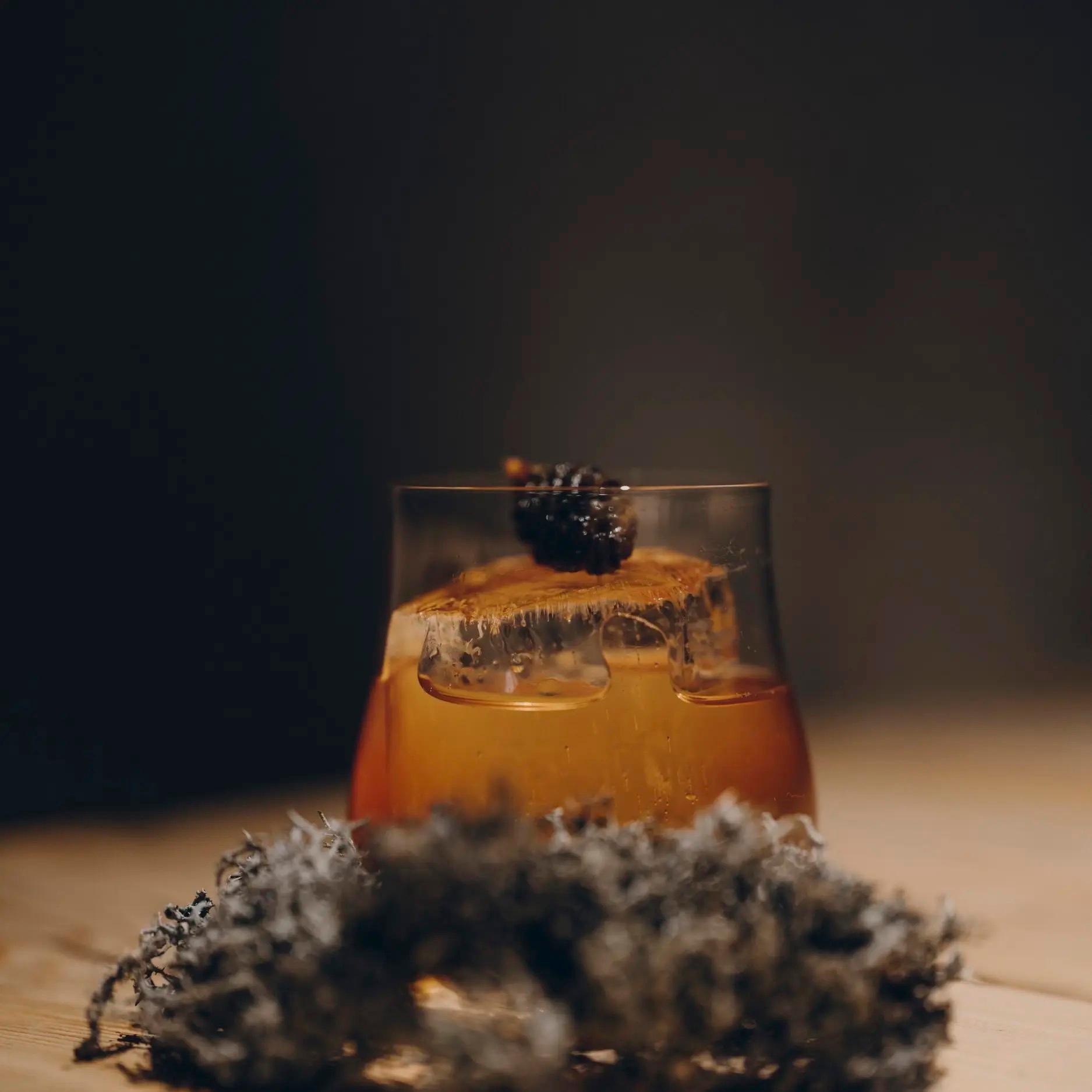 A glass of an amber cocktail with a large ice cube and a berry garnish sits beside a nest of dried moss.