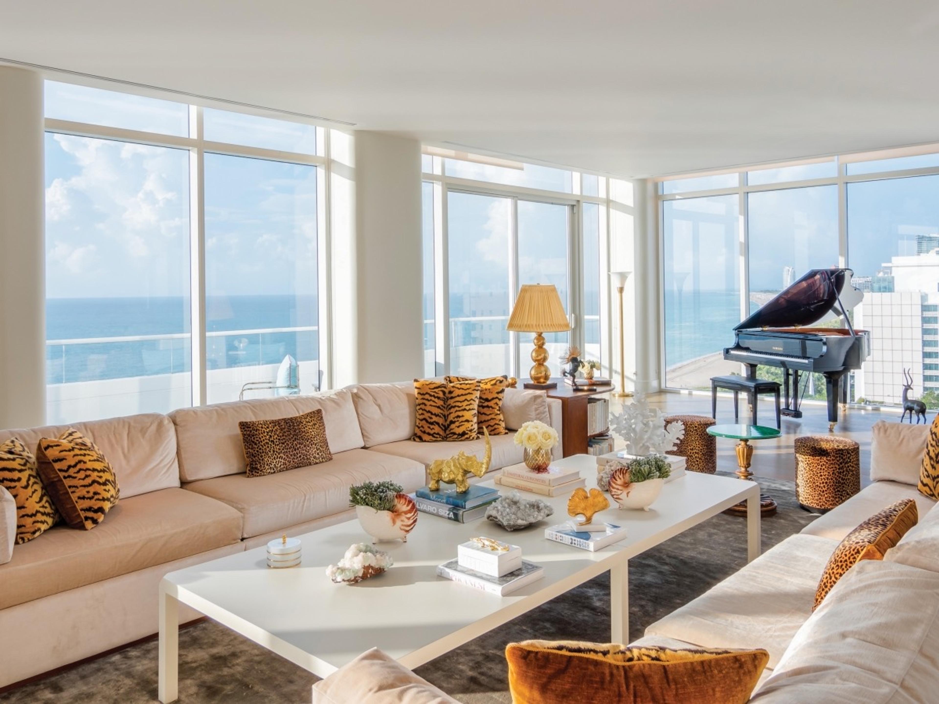 A large residence living room with cream sofas, tiger-print cushions and a grand piano, with floor-to-ceiling windows showing the sea on the left and the city on the right.
