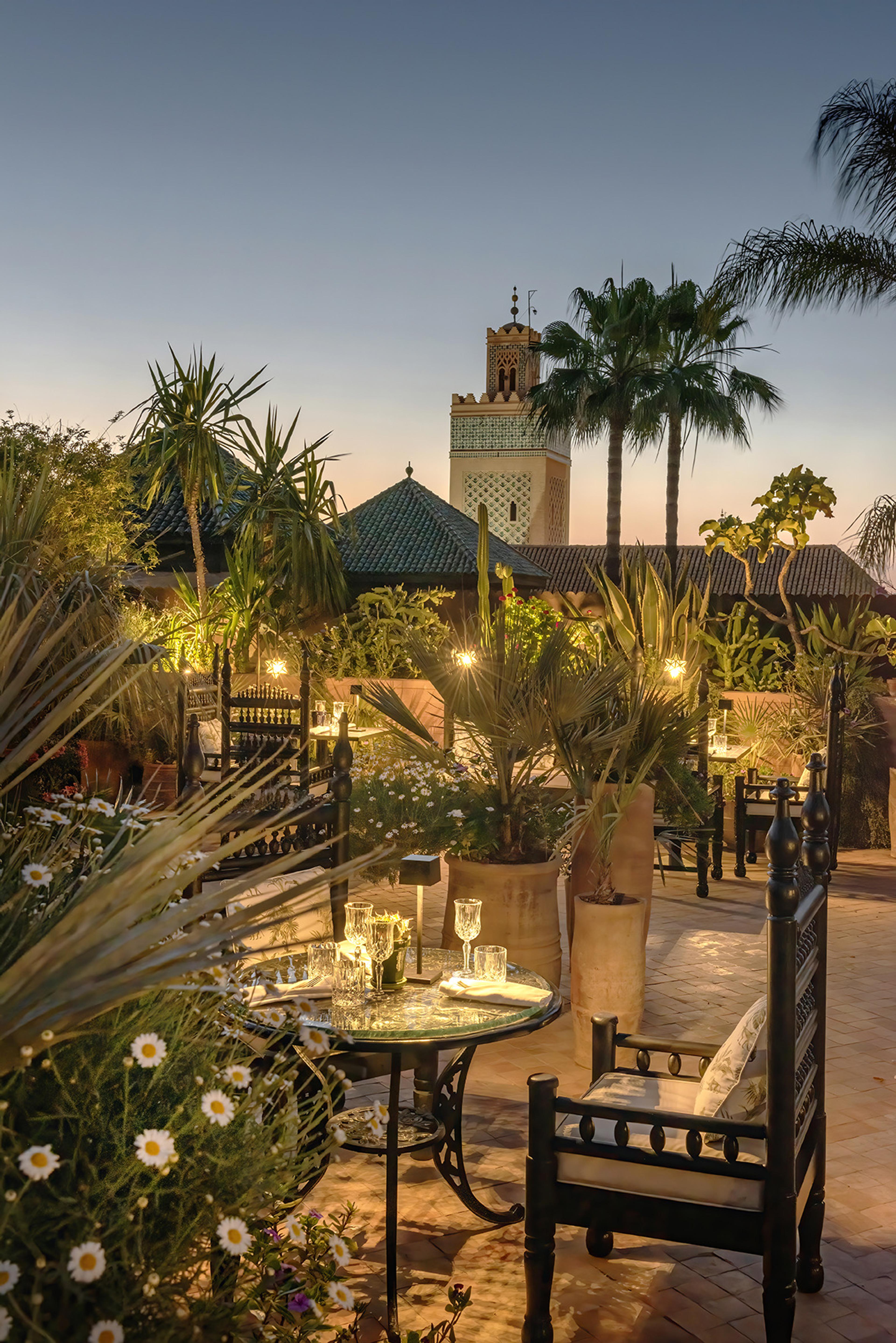 A rooftop terrace set for dinner at dusk, candlelit, with palm trees and a lit minaret in the distance and daisies in the foreground.