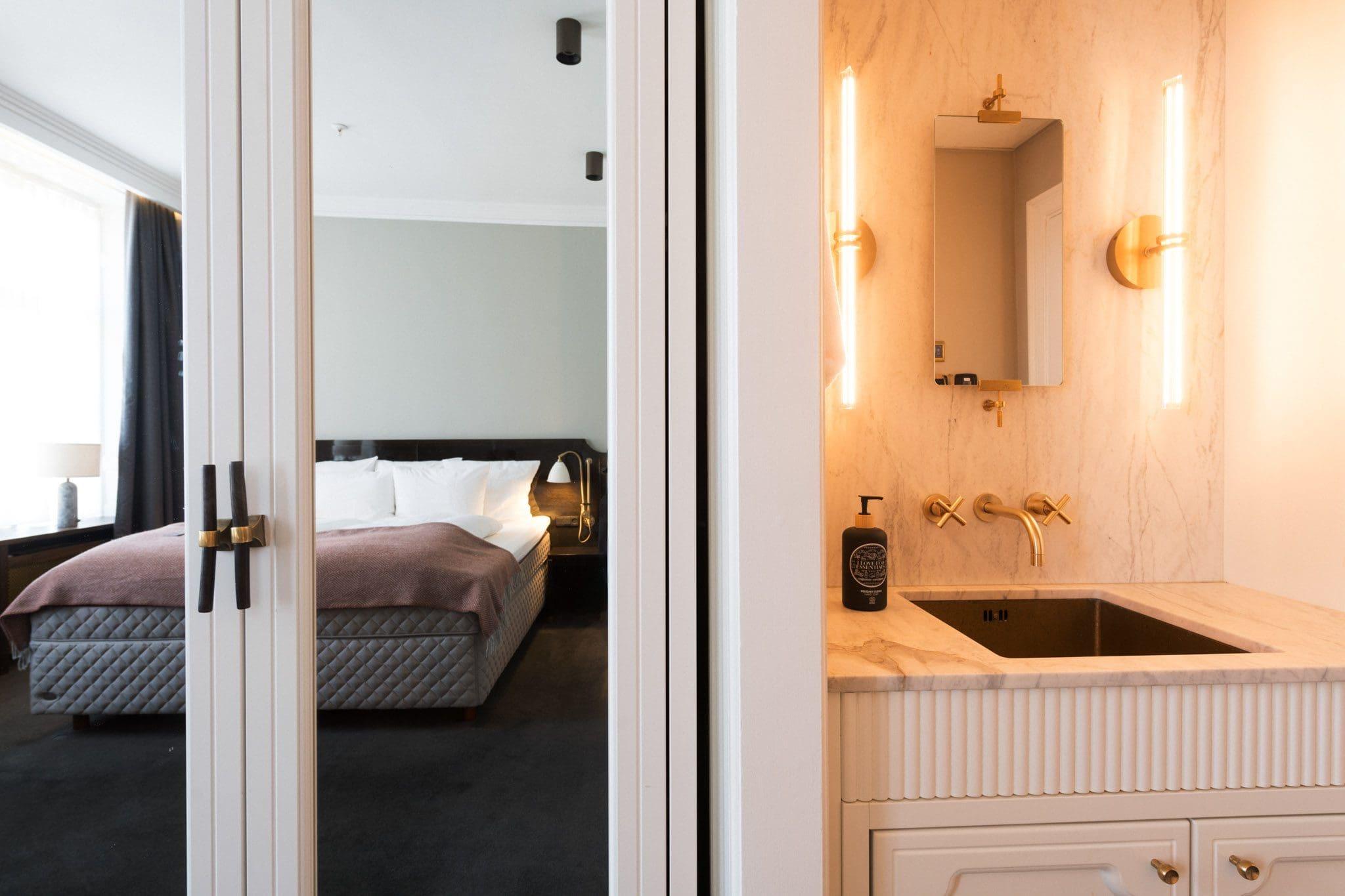 A bathroom vanity with a marble counter, brass taps and wall lights, seen beside an open mirrored door reflecting a bed in the next room.
