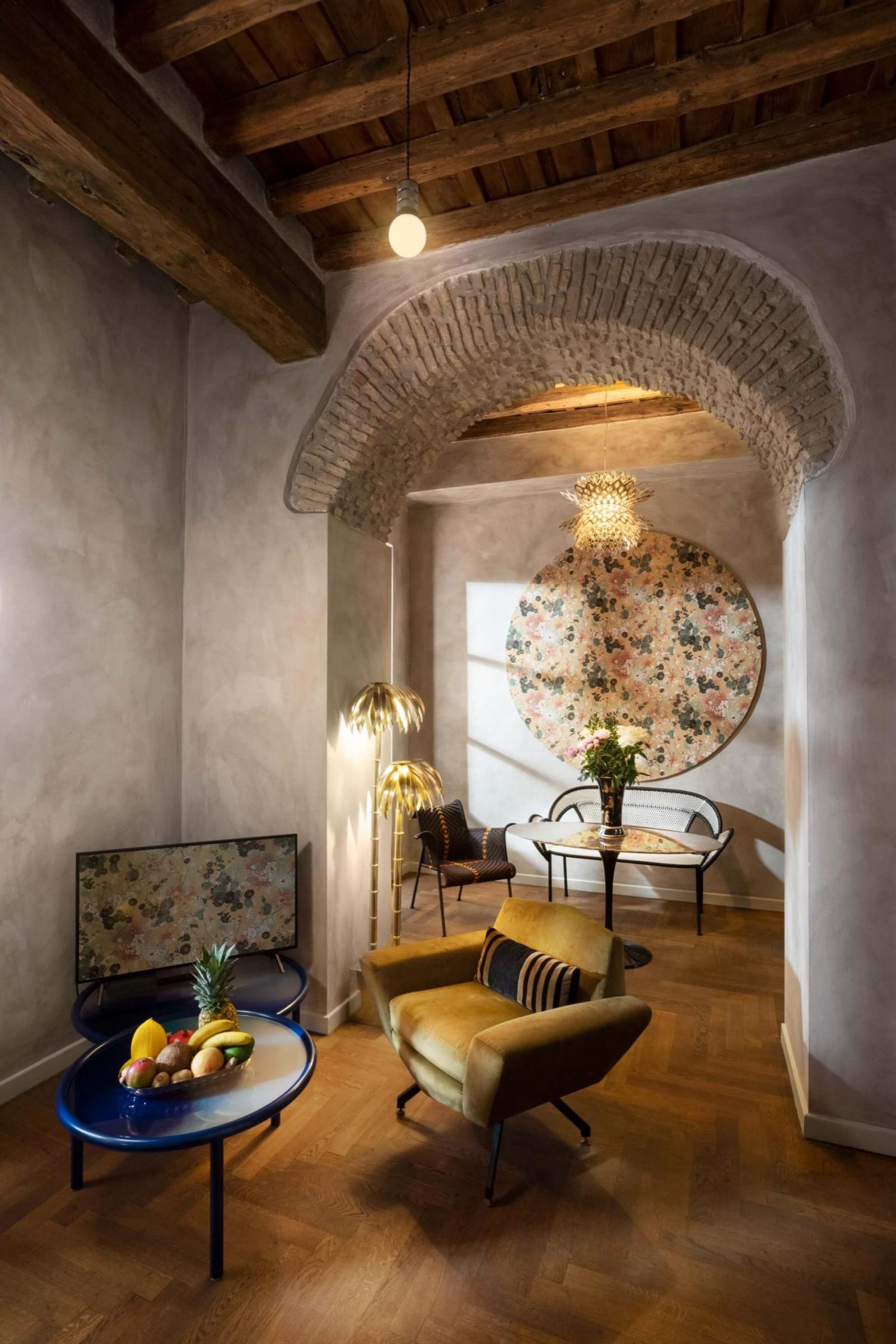 A vaulted room with a brick arch and exposed wooden beams, a blue round coffee table with fruit, a yellow armchair and a round painted panel on the far wall.