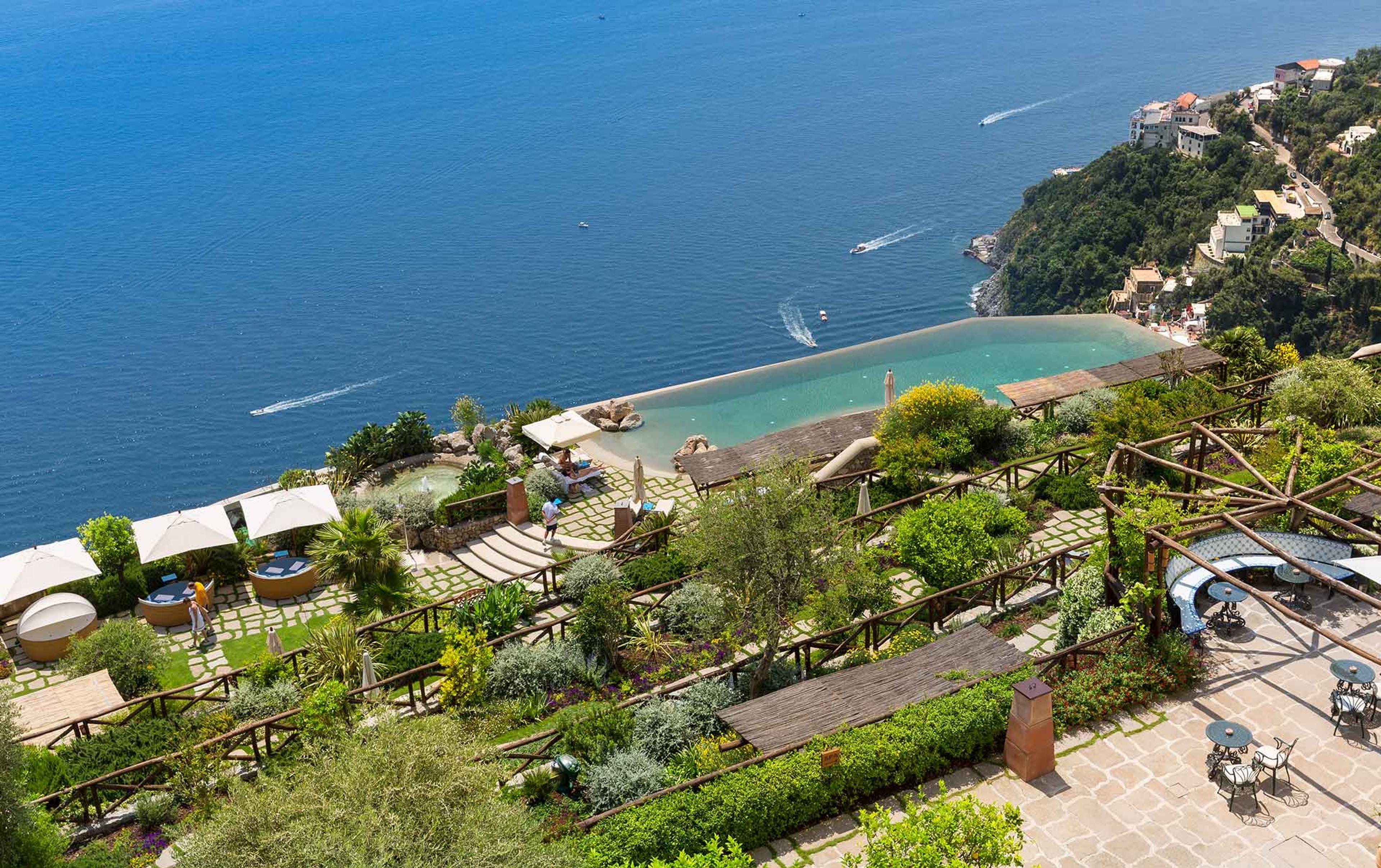 an aerial view of an infinity pool built into the cliff, turquoise sea beyond with two boats, and terraced gardens and seating around it