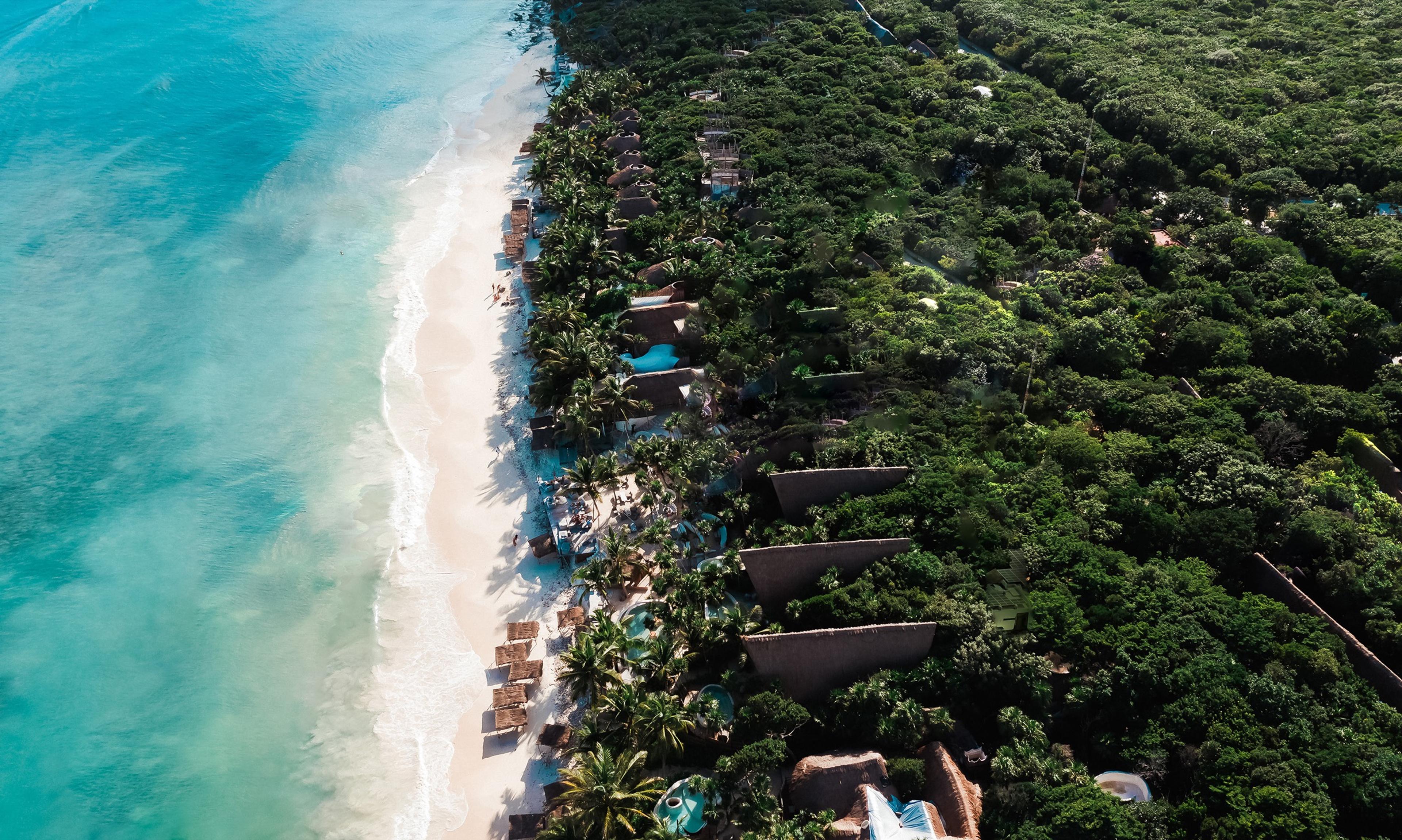 an aerial view of a row of thatched-roof beach casitas and round pools set among trees, beside a white sand beach and turquoise sea