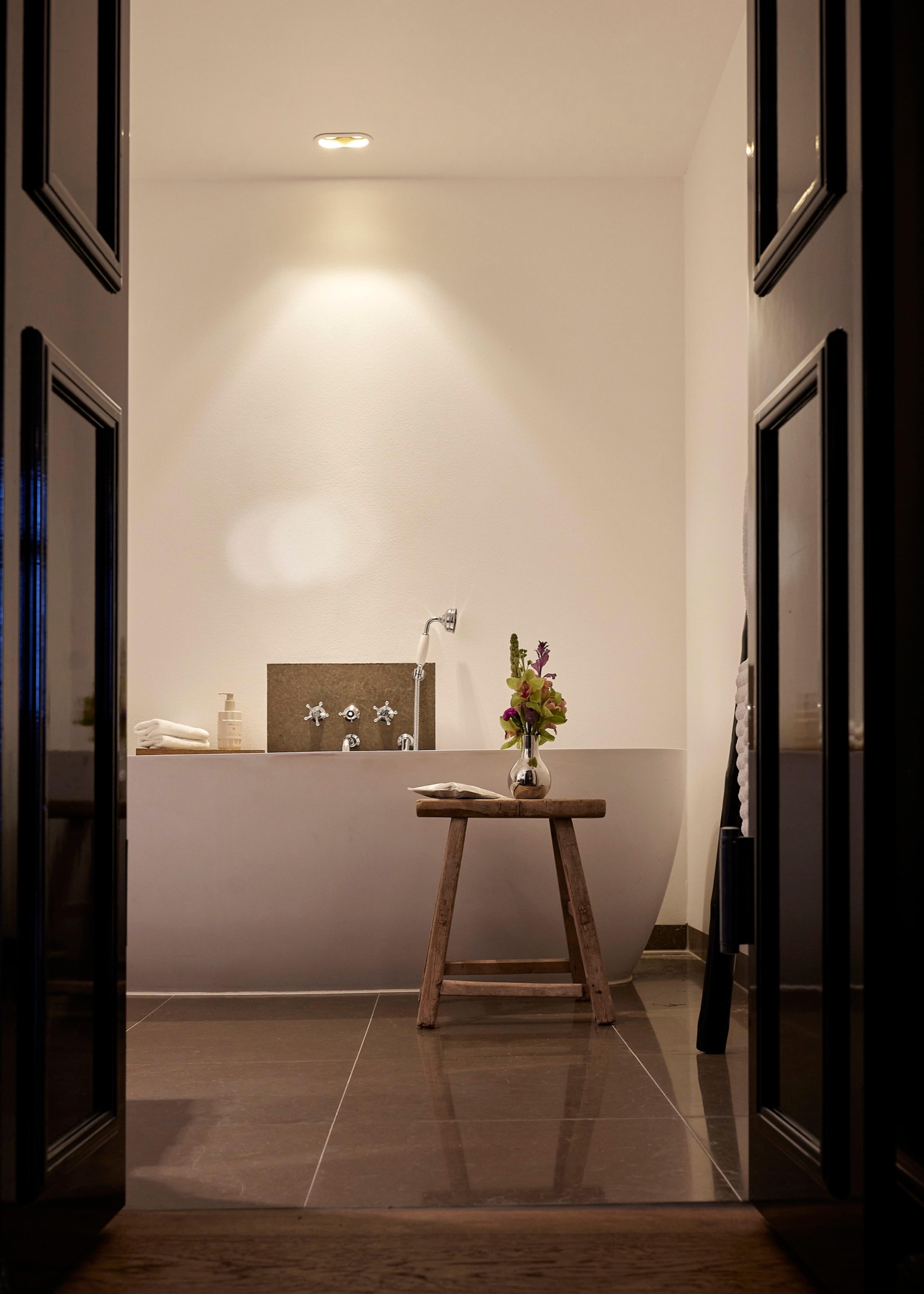 A freestanding bathtub with wall-mounted taps stands behind a wooden stool holding a small vase of flowers, seen through an open doorway.