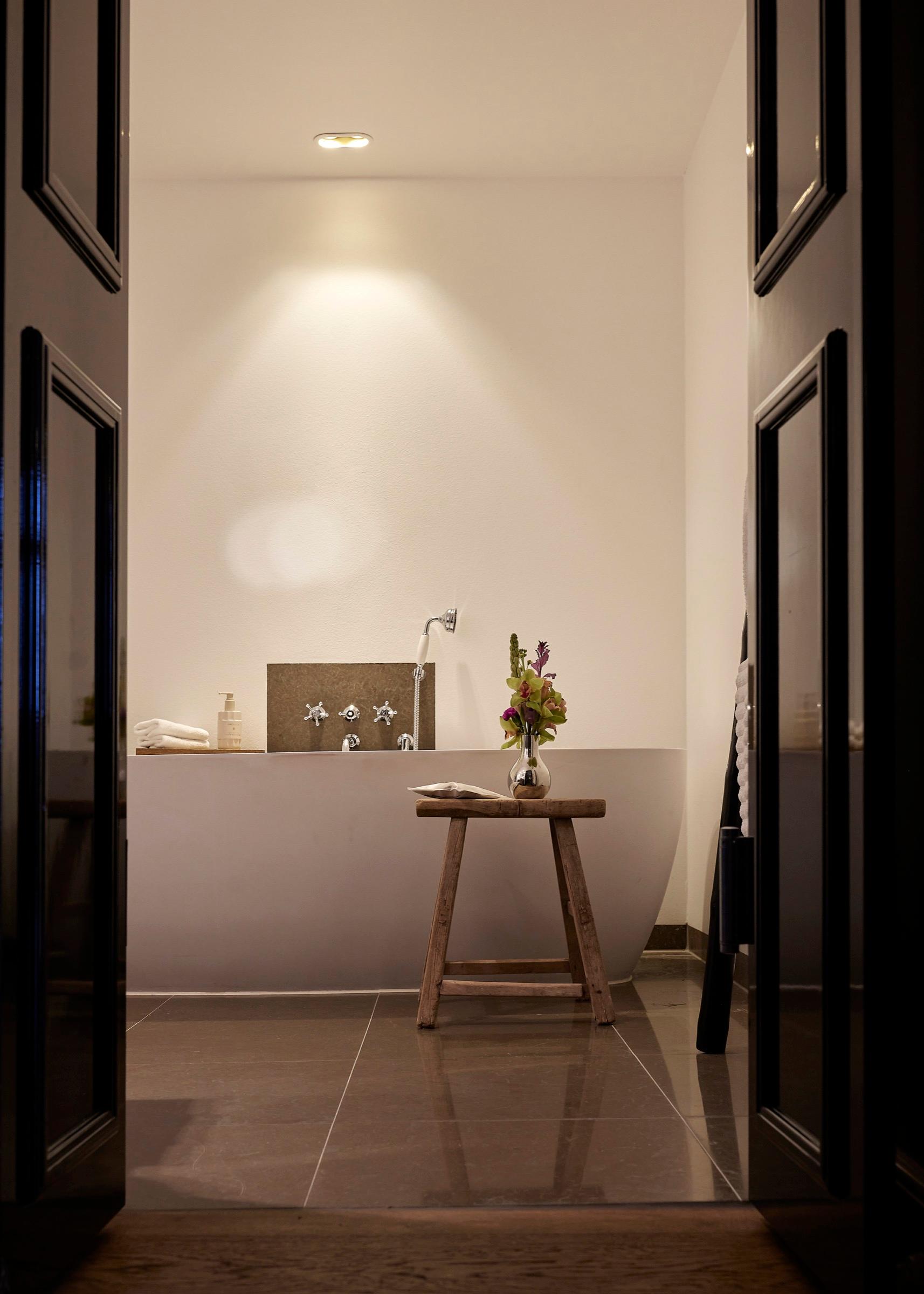 A freestanding bathtub with wall-mounted taps stands behind a wooden stool holding a small vase of flowers, seen through an open doorway.
