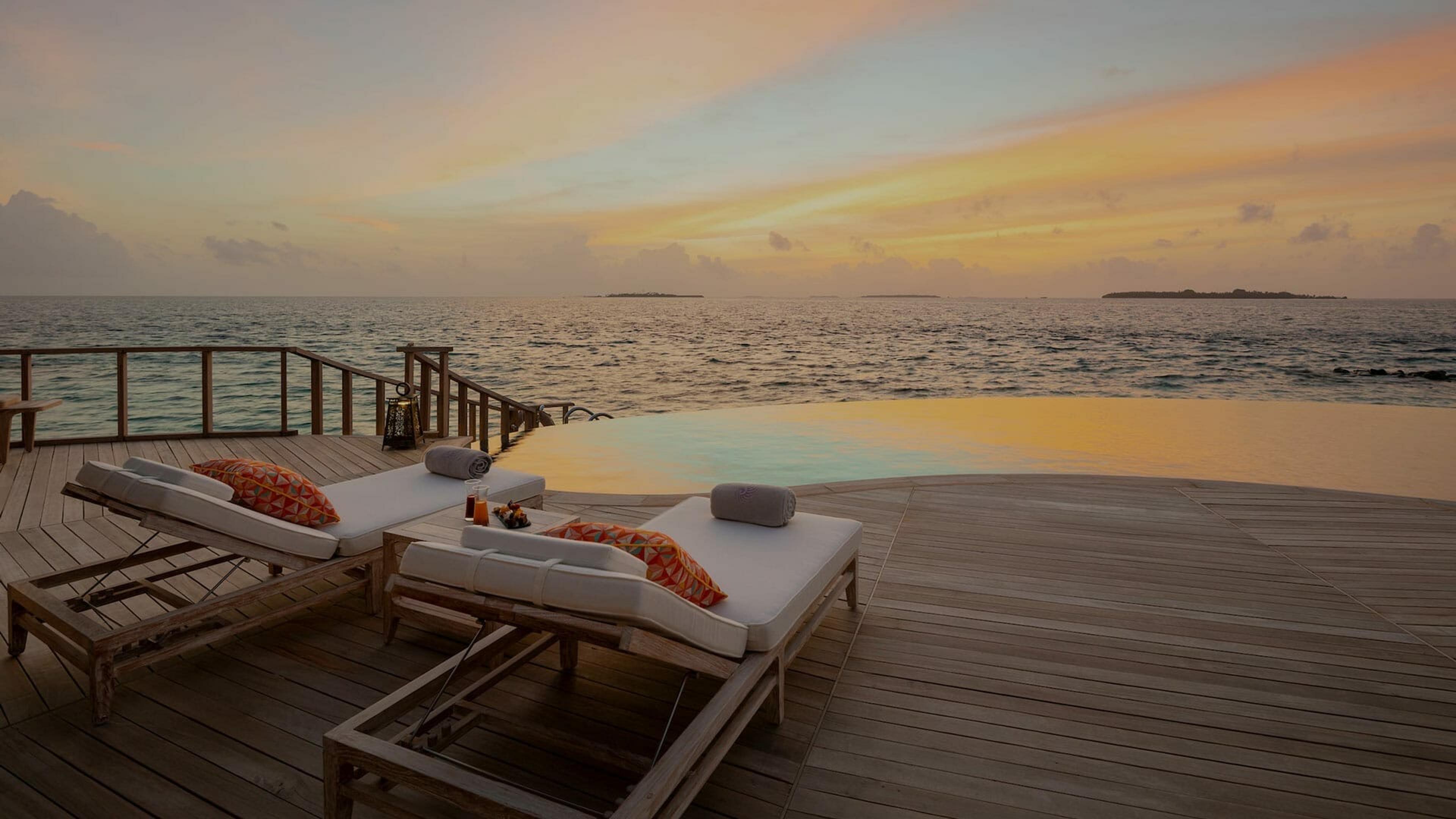 Two empty sun loungers on a wooden deck beside an infinity pool at sunset, with small islands visible on the horizon.