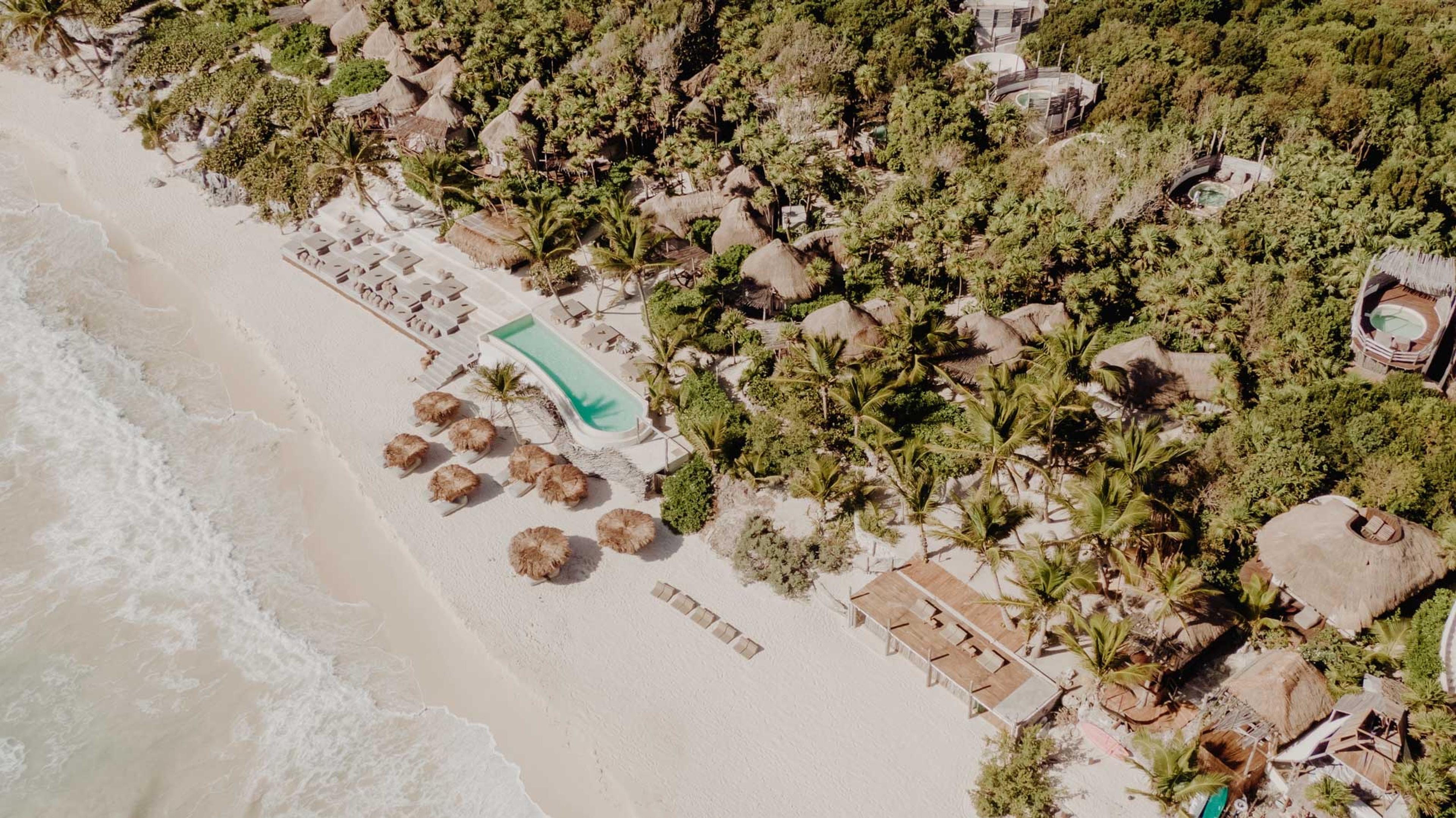 an aerial view of a long pool and a row of palm-thatched sun shades on a white sand beach, with villas in the jungle beyond