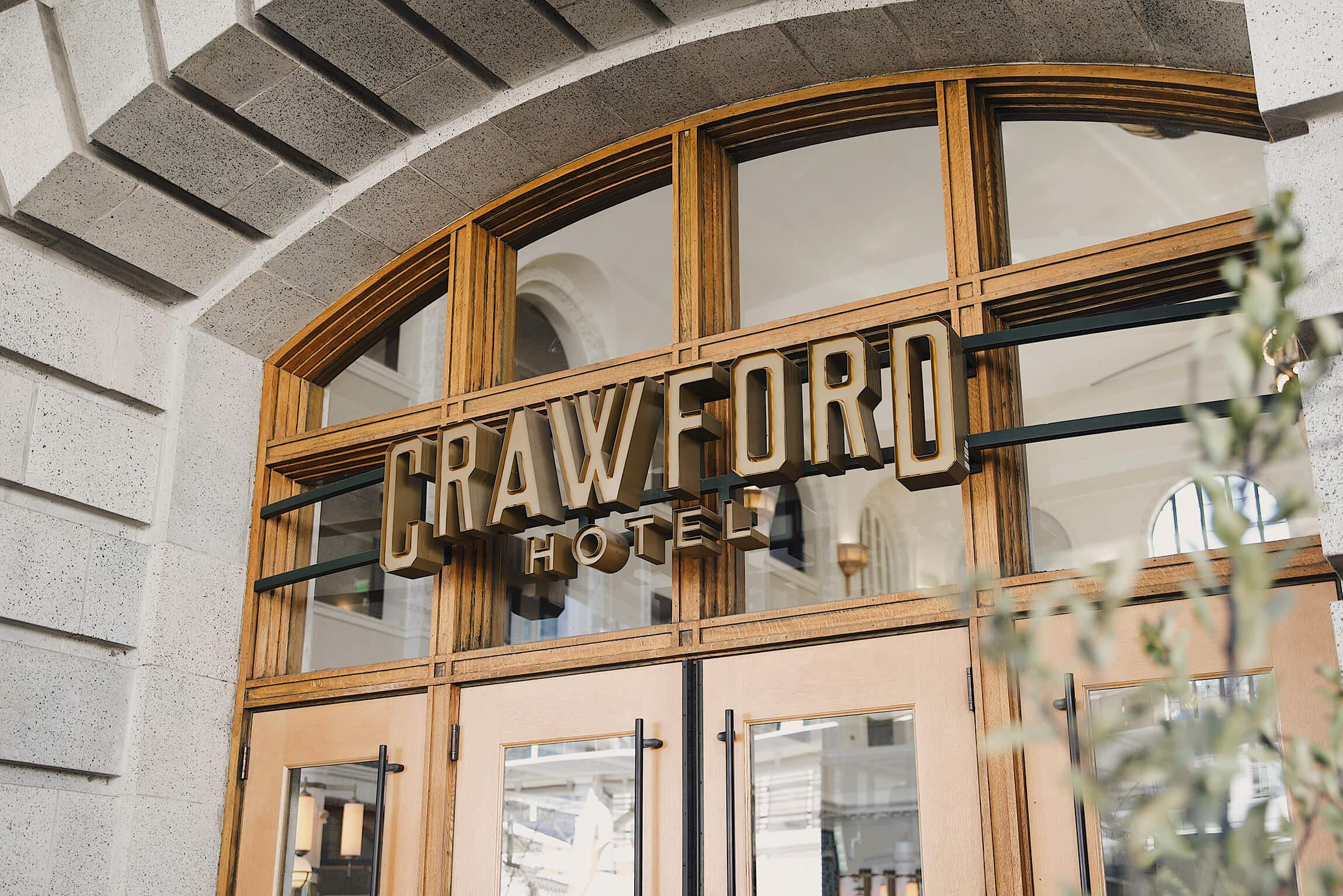Brass lettering reading CRAWFORD HOTEL above glass doors set in a stone arch, with a branch in the foreground.