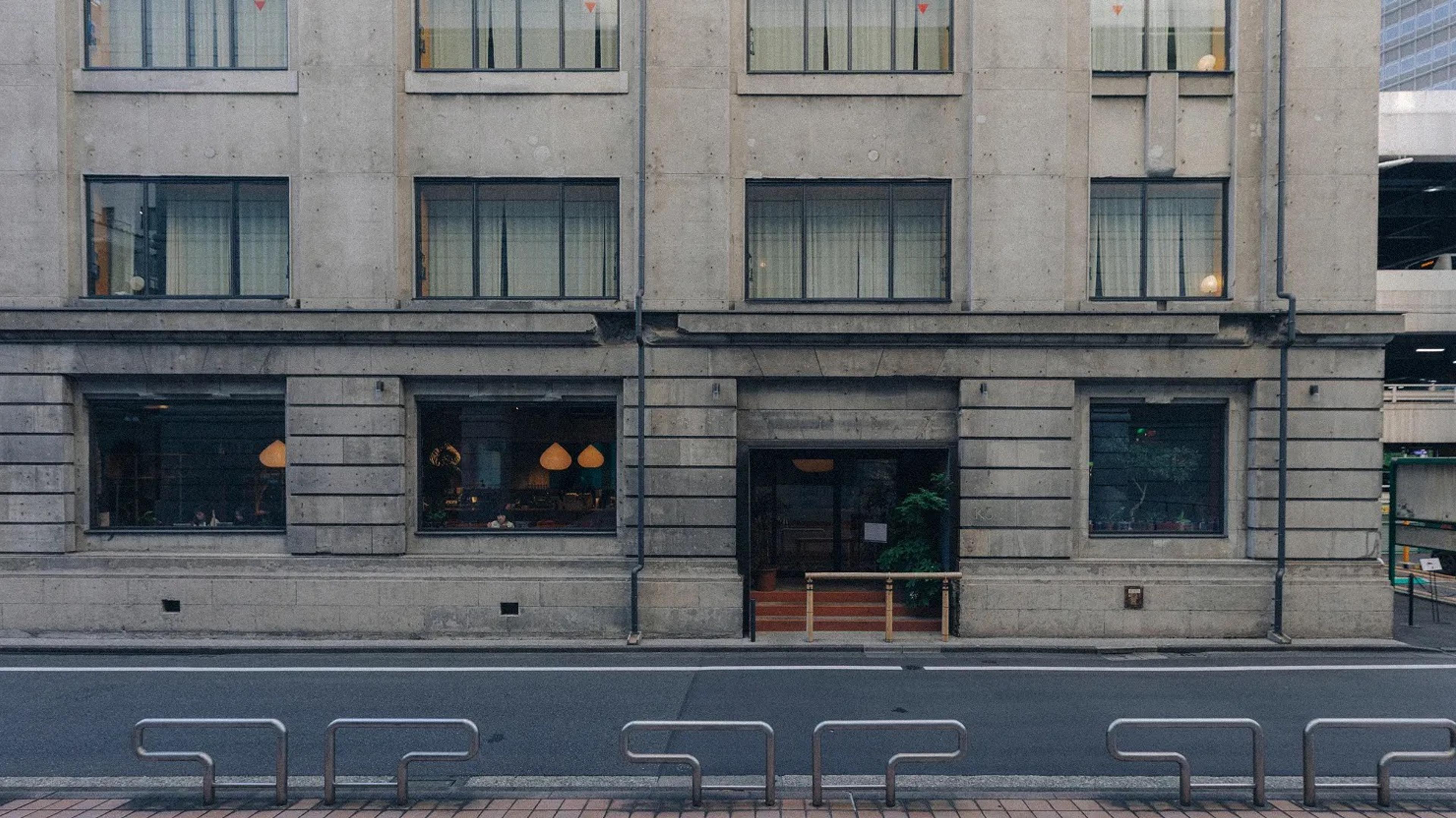 A concrete hotel building with three rows of windows above street level, a recessed glass entrance up a few steps between two lit shopfront windows, and bicycle racks lined along the pavement out front.