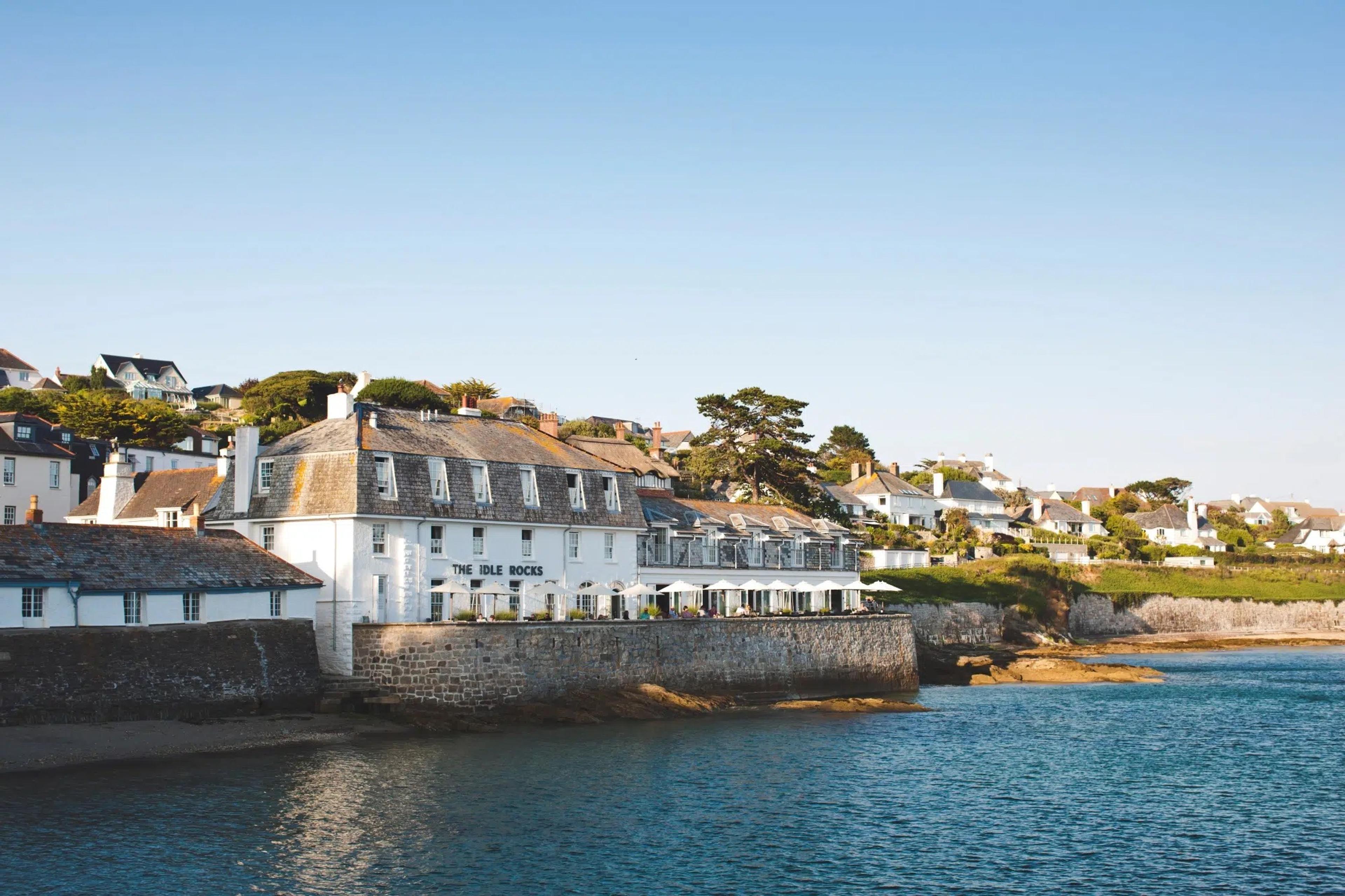 A white harbourside hotel building behind a stone sea wall, with umbrellas along its terrace, houses on a wooded hillside behind, and calm blue water in front, lit by evening sun.