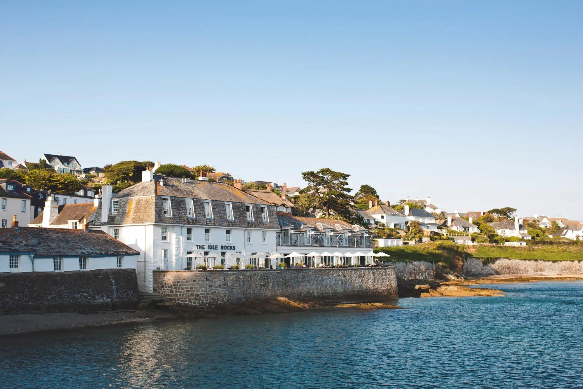 A white harbourside hotel building behind a stone sea wall, with umbrellas along its terrace, houses on a wooded hillside behind, and calm blue water in front, lit by evening sun.