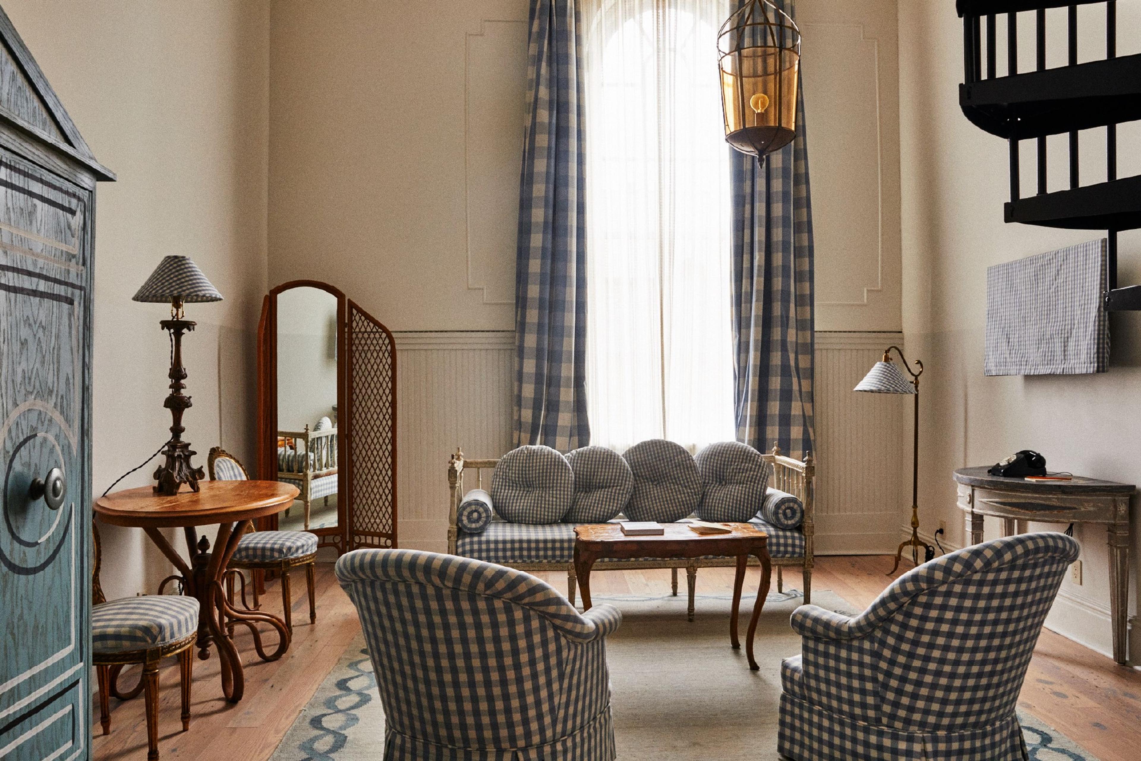 A high-ceilinged sitting room with two checked armchairs and a matching bench sofa around a low table, tall curtained windows, a wall-mounted spiral stair, and a painted armoire.