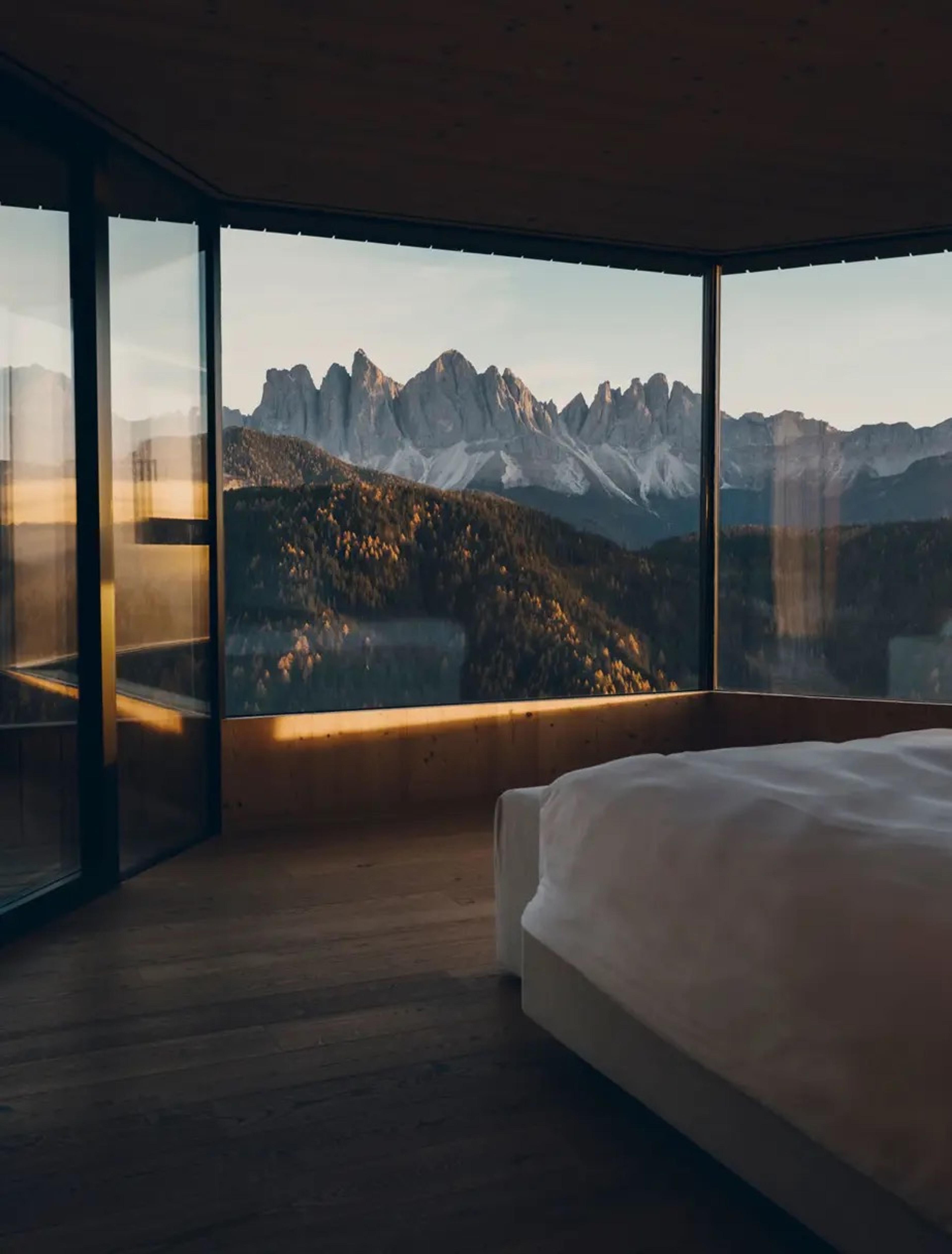 A bedroom with floor-to-ceiling windows opens onto a terrace facing jagged mountain peaks at dusk.