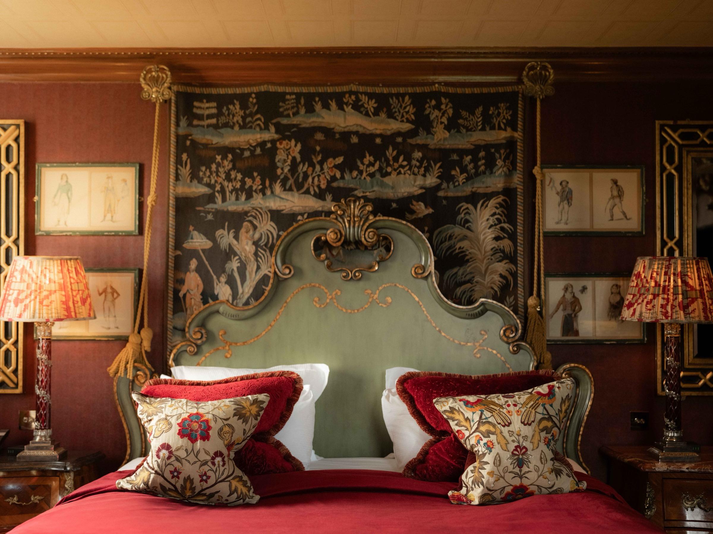 a bed with an ornate pale green upholstered headboard, a tapestry above it and patterned cushions on a red throw