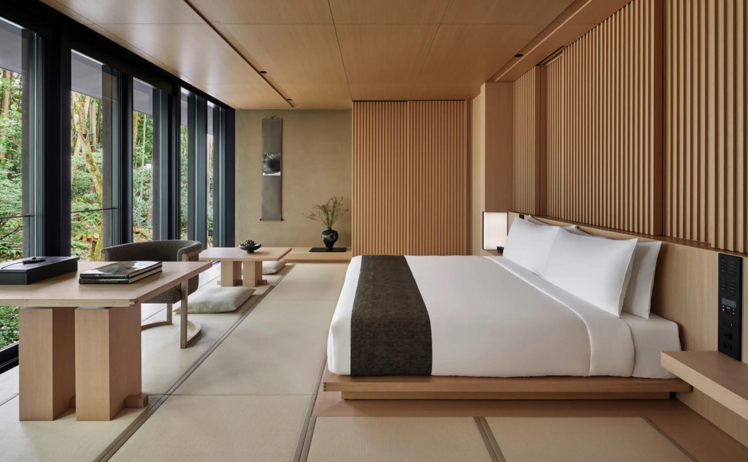 A bedroom with a platform bed dressed in white linen and a dark throw, a desk and chair by floor-to-ceiling windows, and a scroll on the wall.