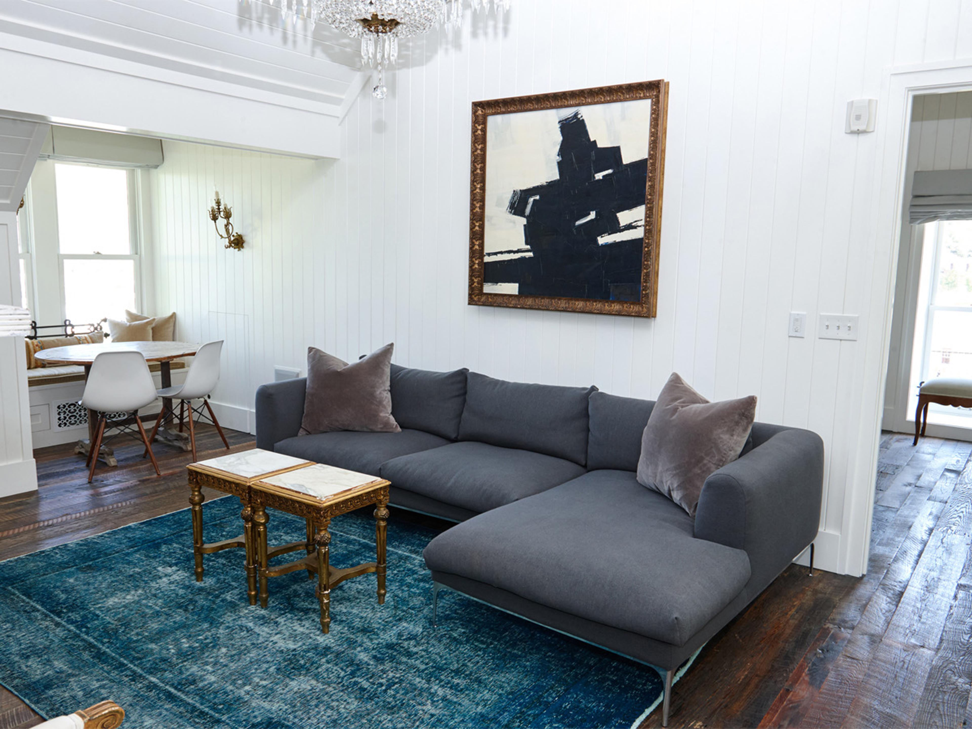 A grey sectional sofa with velvet cushions facing a large abstract painting, with a breakfast nook and chandelier visible beyond.