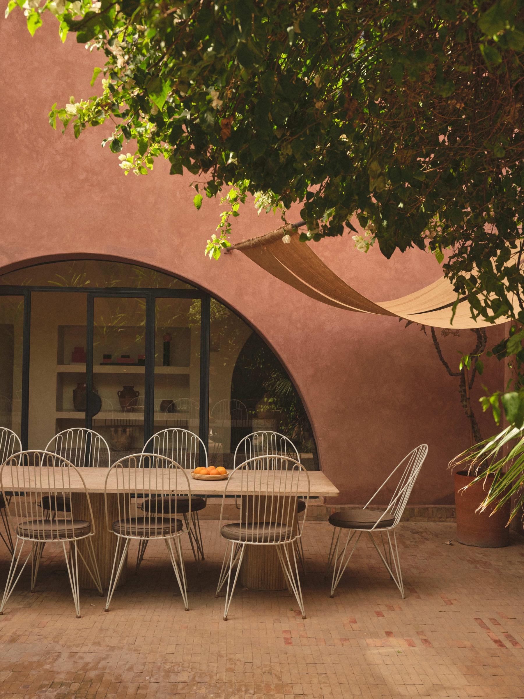 an outdoor dining table set with eight wire chairs and a bowl of oranges, under a woven hammock hanging from a tree, in front of an arched window