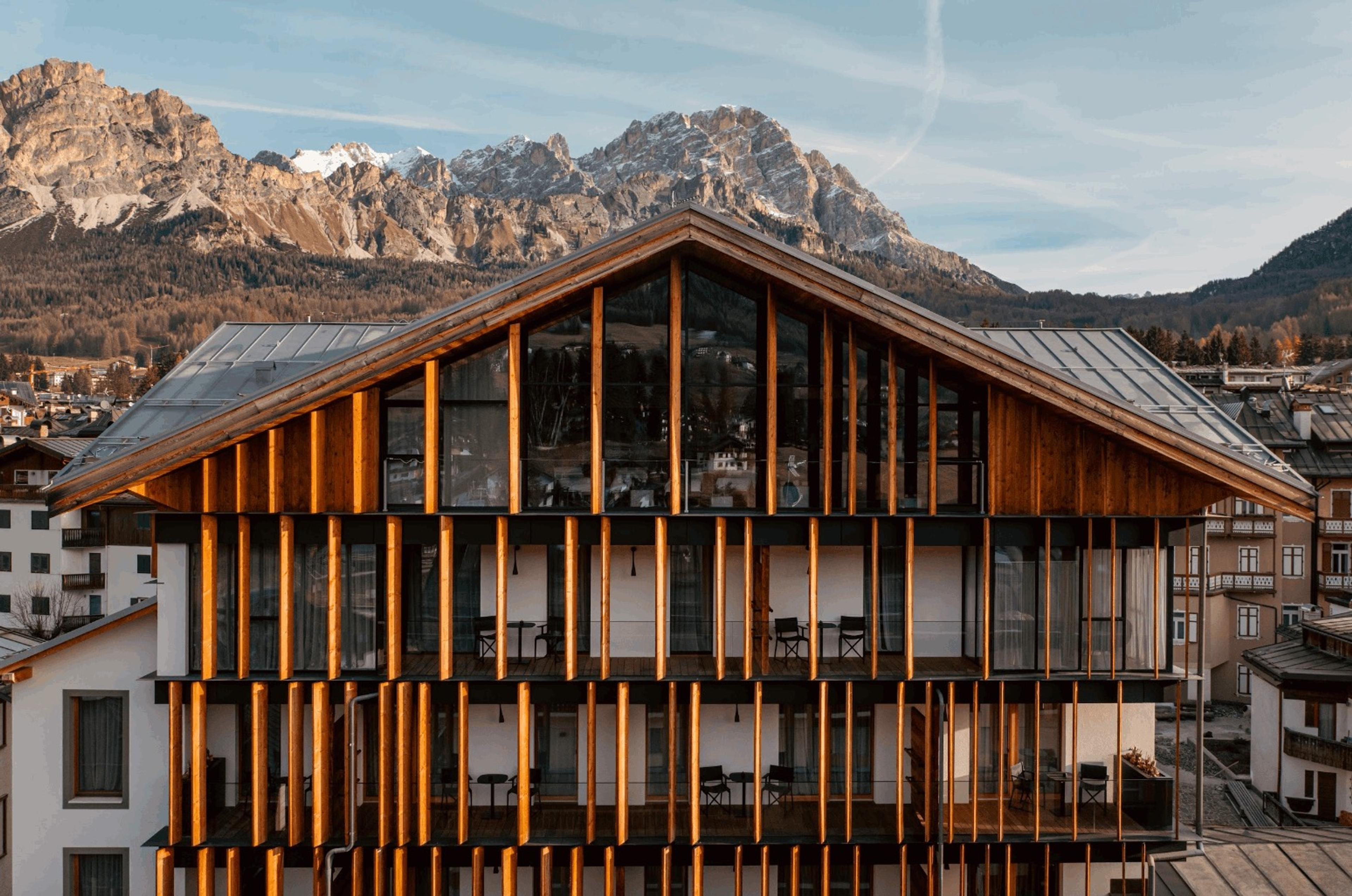 The hotel's timber-clad facade rises against snow-capped mountain peaks lit by evening sun, with balconies along each floor.