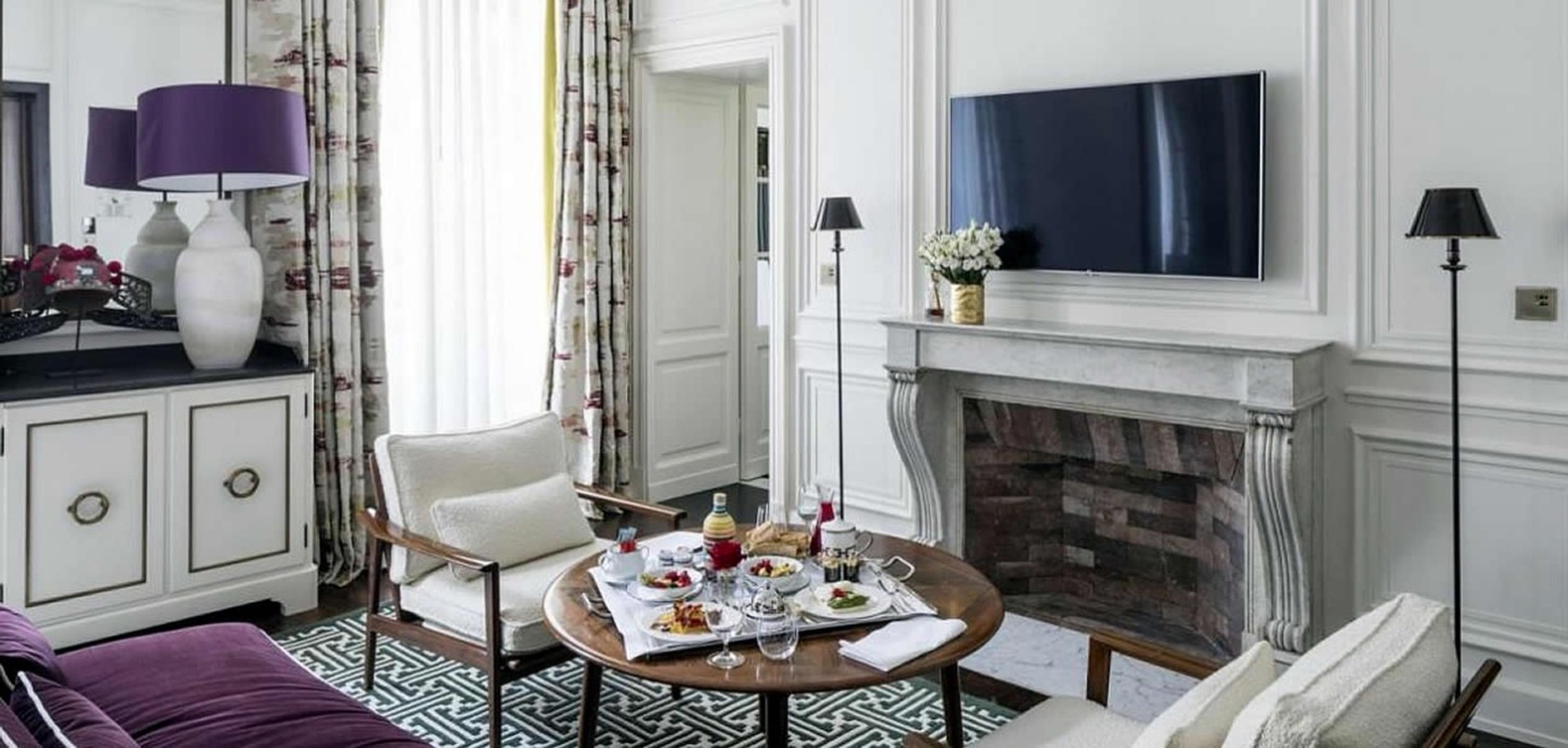 A breakfast tray of pastries, yoghurt and juice set on a round table in a sitting room, with a television above a marble fireplace.