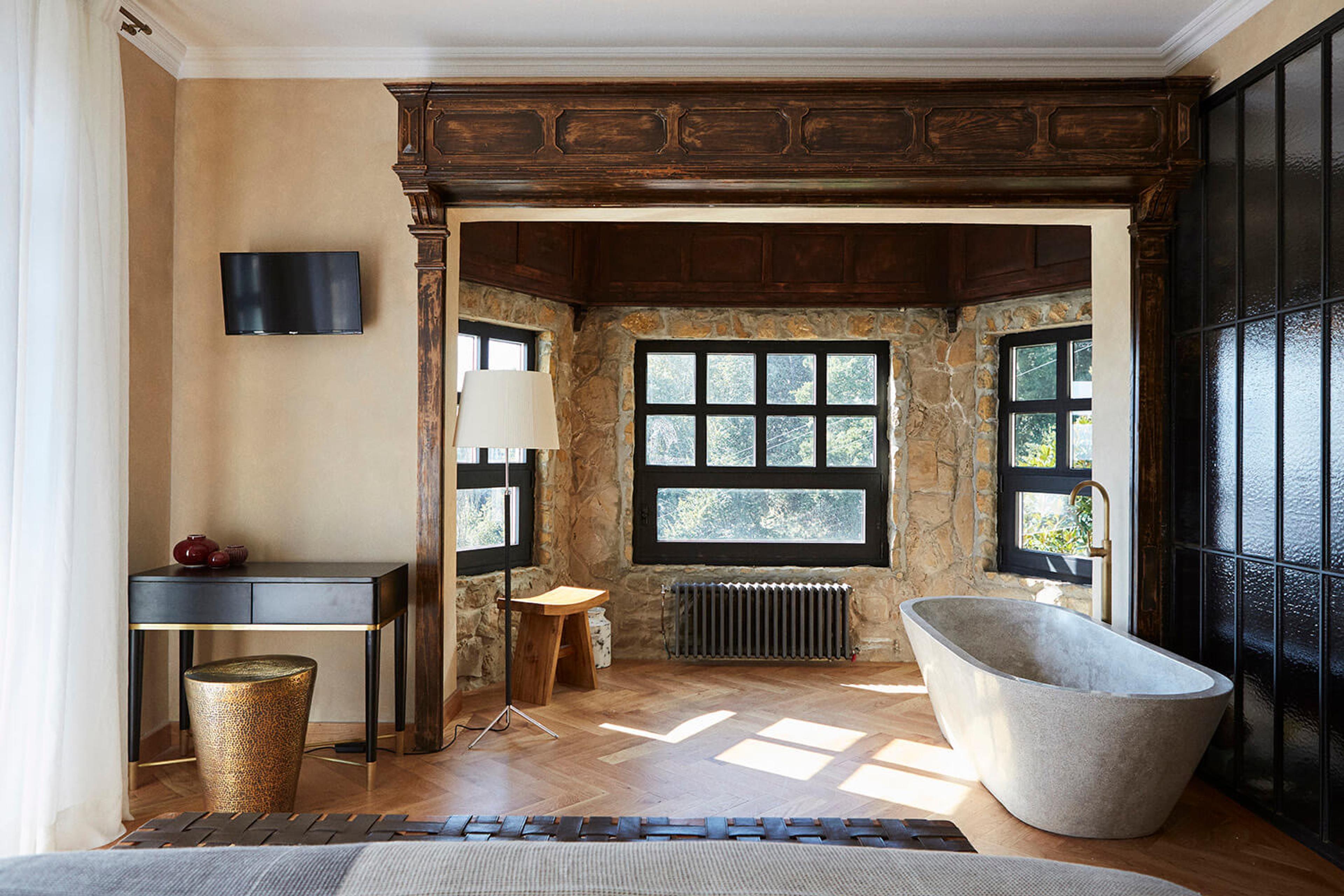 a freestanding stone bathtub in a bathroom with a carved wood beam over stone-walled windows, a wall-mounted television and a wooden stool