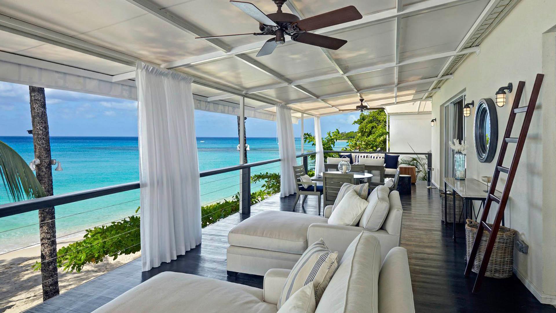 A covered terrace with white cushioned daybeds and dining chairs looking straight out over turquoise sea.