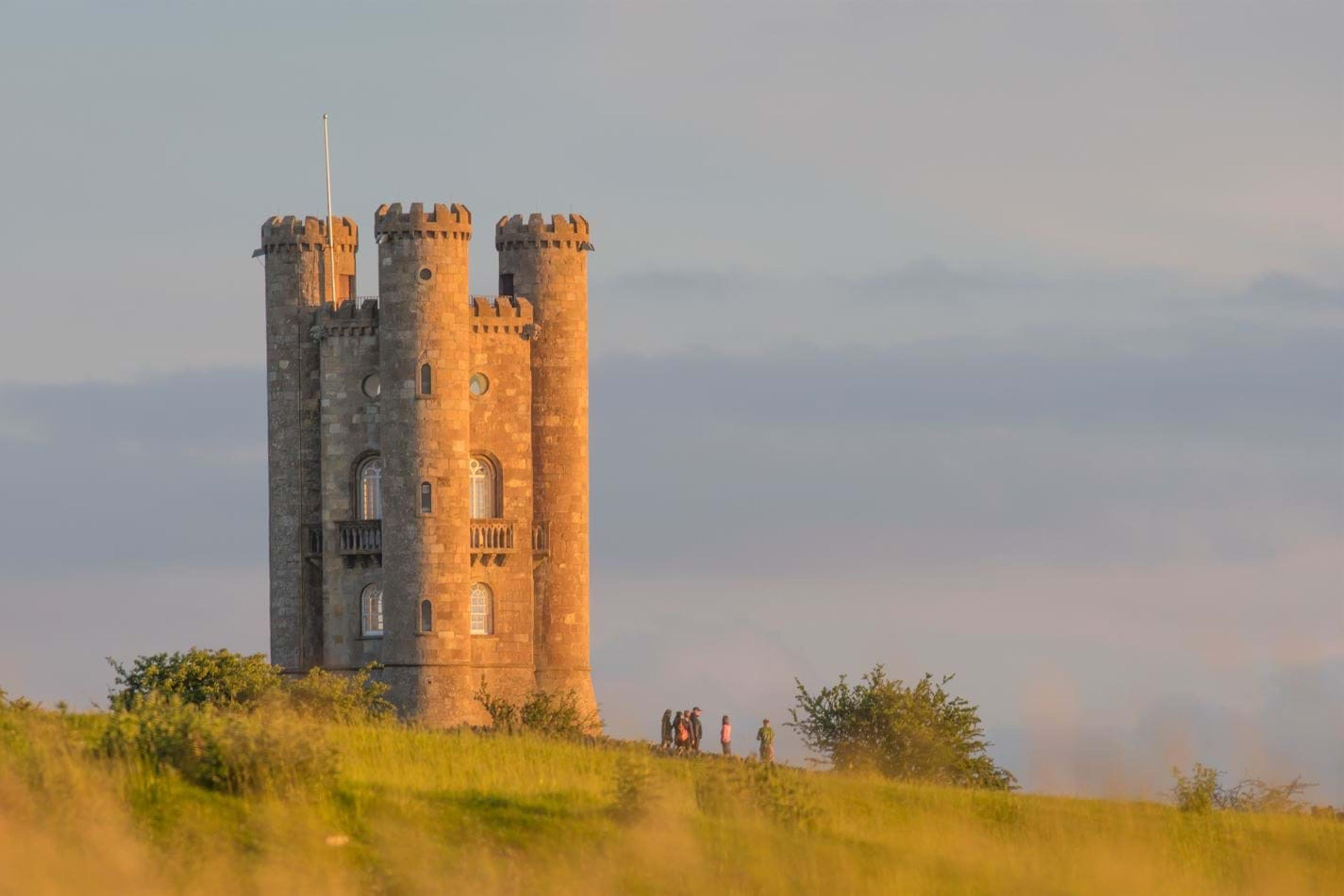 Broadway Tower stands on a grassy hilltop at golden hour, with a small group of walkers at its base.