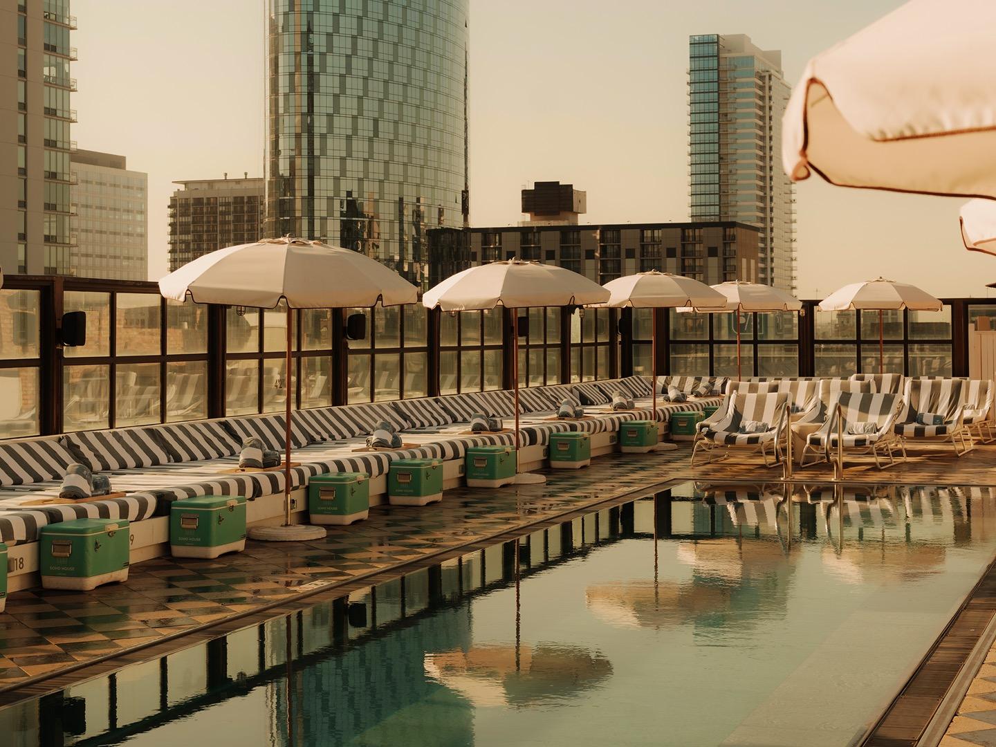 A rooftop pool lined with striped day beds and green ice boxes under white umbrellas, with the city skyline behind it at dusk.