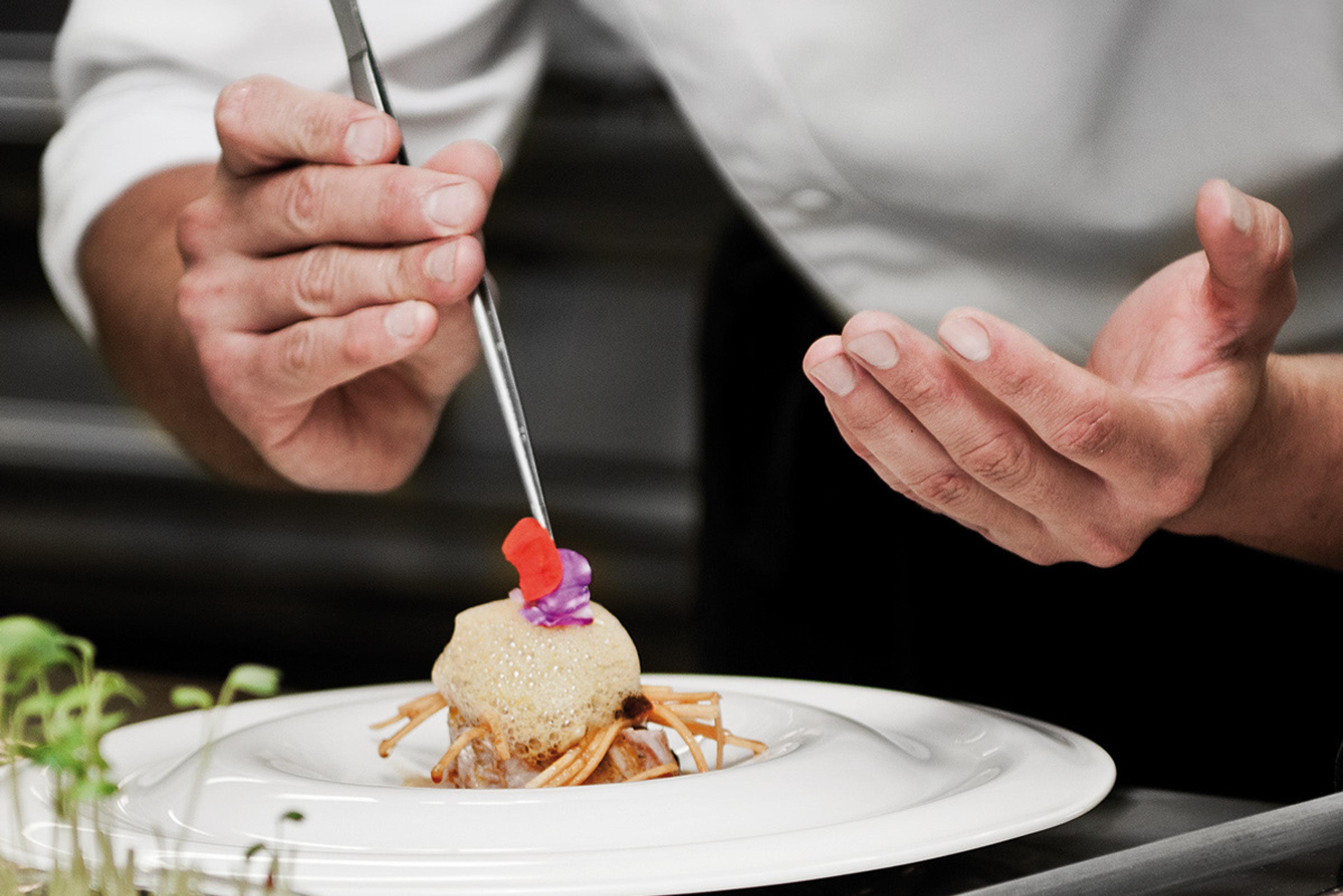 A chef's hands plating a dessert with tweezers, placing a small garnish on a pale sphere set in a white bowl.