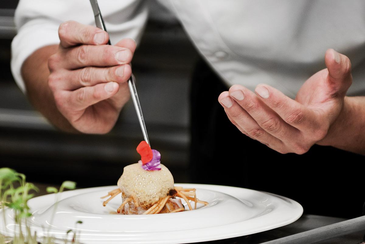 A chef's hands plating a dessert with tweezers, placing a small garnish on a pale sphere set in a white bowl.