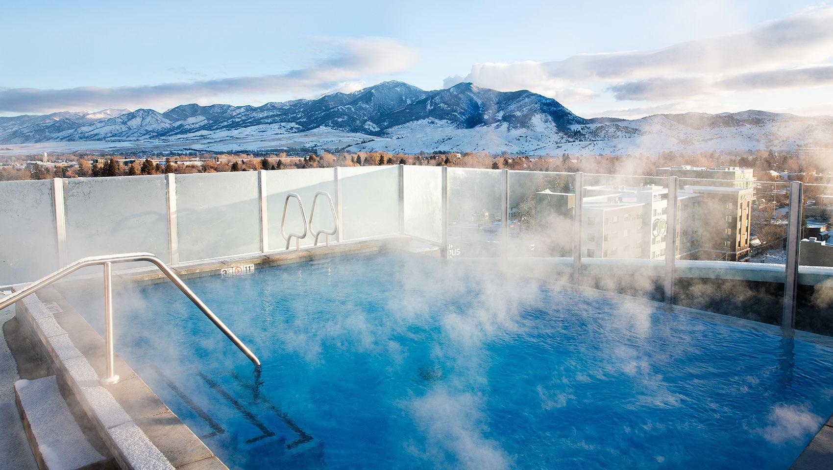 A steaming rooftop pool with a metal handrail at its steps, snow-covered mountains and the town below under a pale sky.
