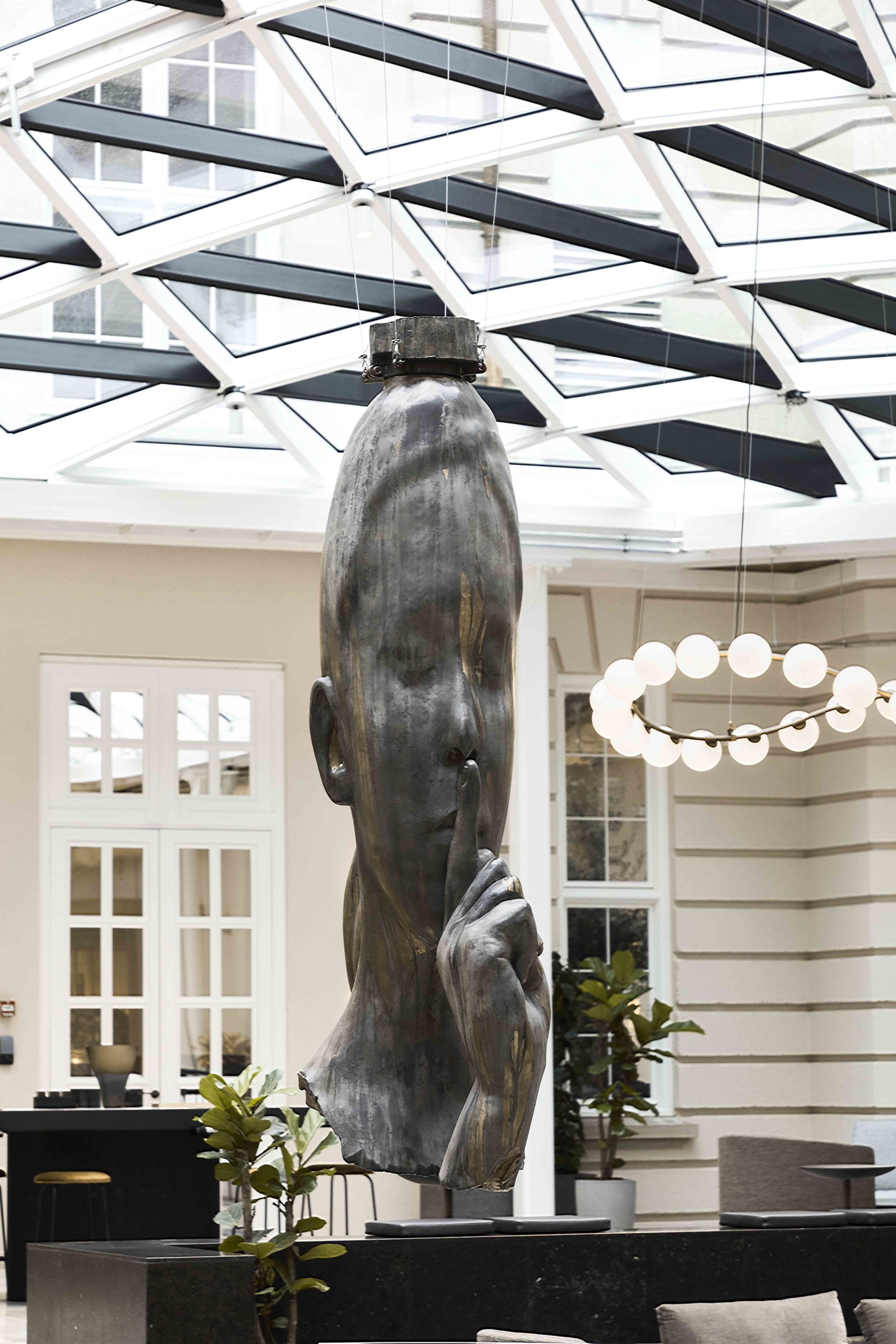 A glass-roofed atrium with a large bronze sculpture of a face suspended overhead, a ring of globe lights, potted plants and seating below.