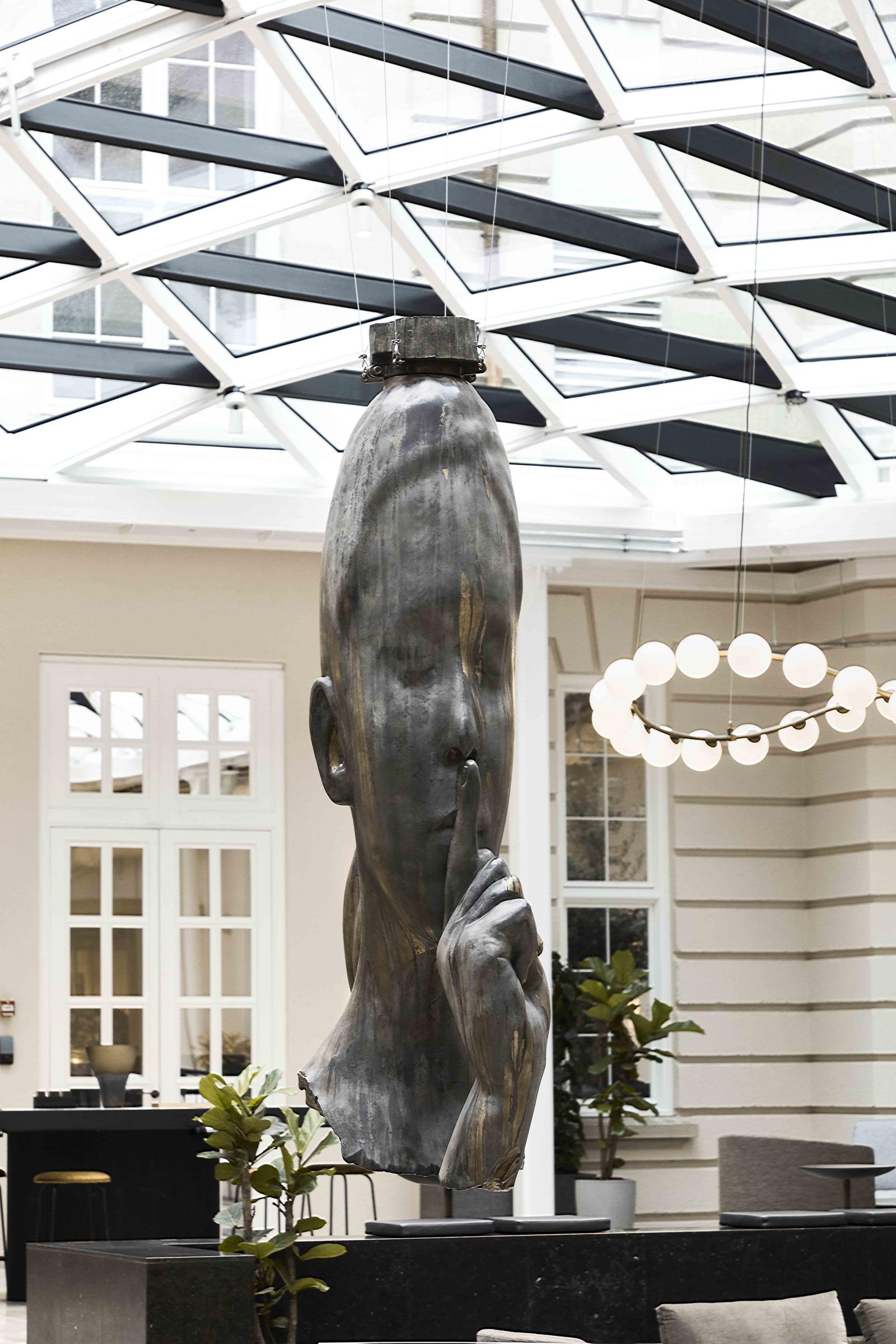 A glass-roofed atrium with a large bronze sculpture of a face suspended overhead, a ring of globe lights, potted plants and seating below.