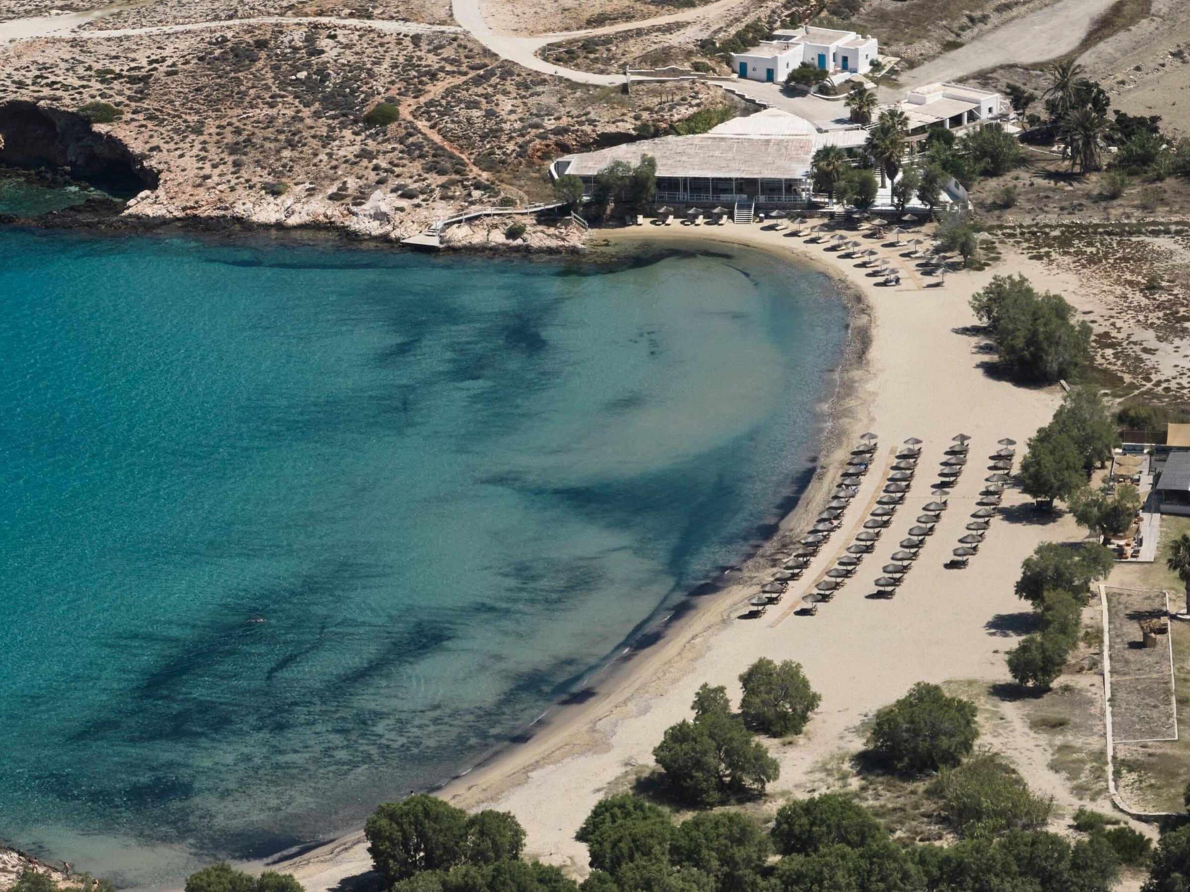A curved sandy cove of turquoise water on the west coast of Paros, with rows of umbrellas along the beach and low white buildings above it